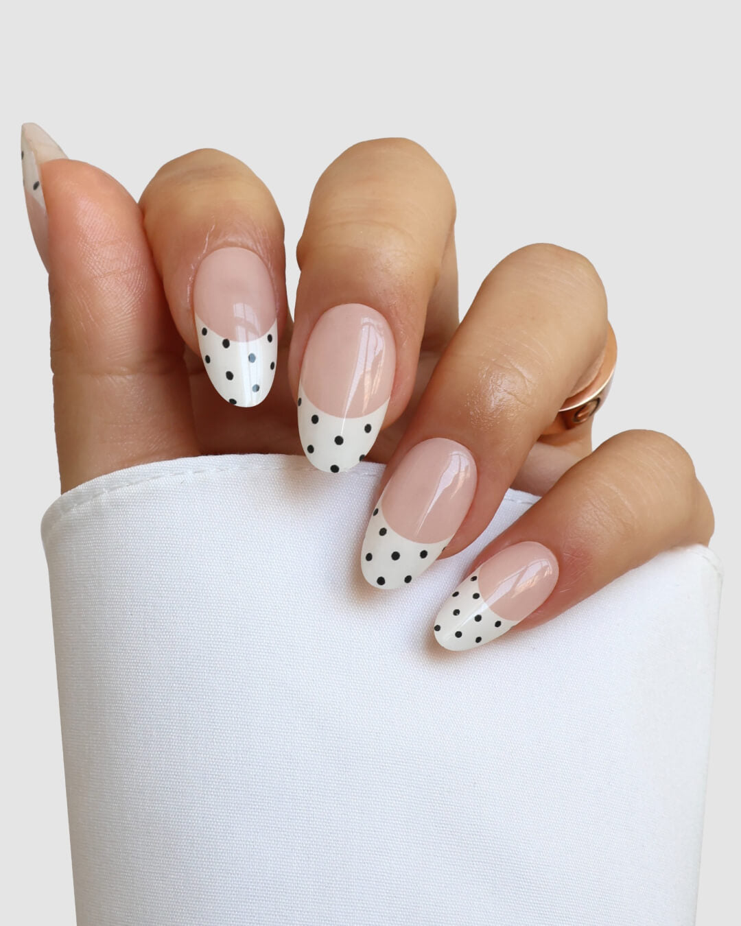 A hand showcases Sassy Saints - Brings The Salon Home's French with Benefits Gel Wrap: almond-shaped nails in a French manicure with white tips and black polka dots, against a light background. The person wears a white sleeve.