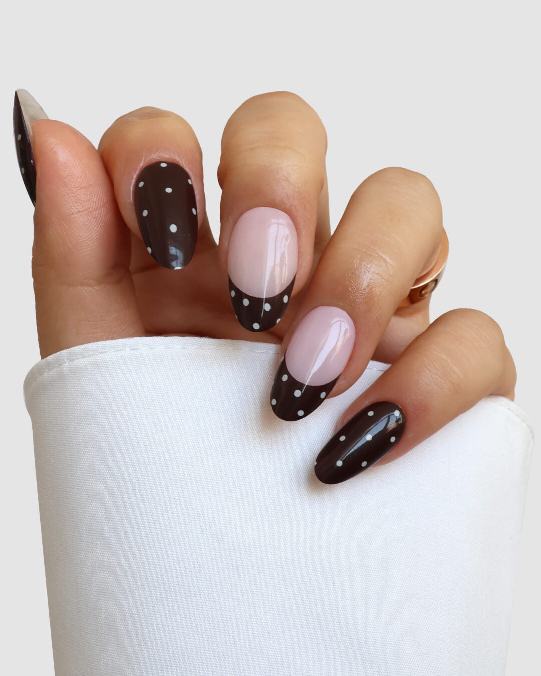 A hand with almond-shaped nails sports Sassy Saints' "Dark Side Darling Gel Wrap"—a chic French manicure design with black tips, white polka dots, and natural pink bases. The look is partially concealed by a white sleeve.
