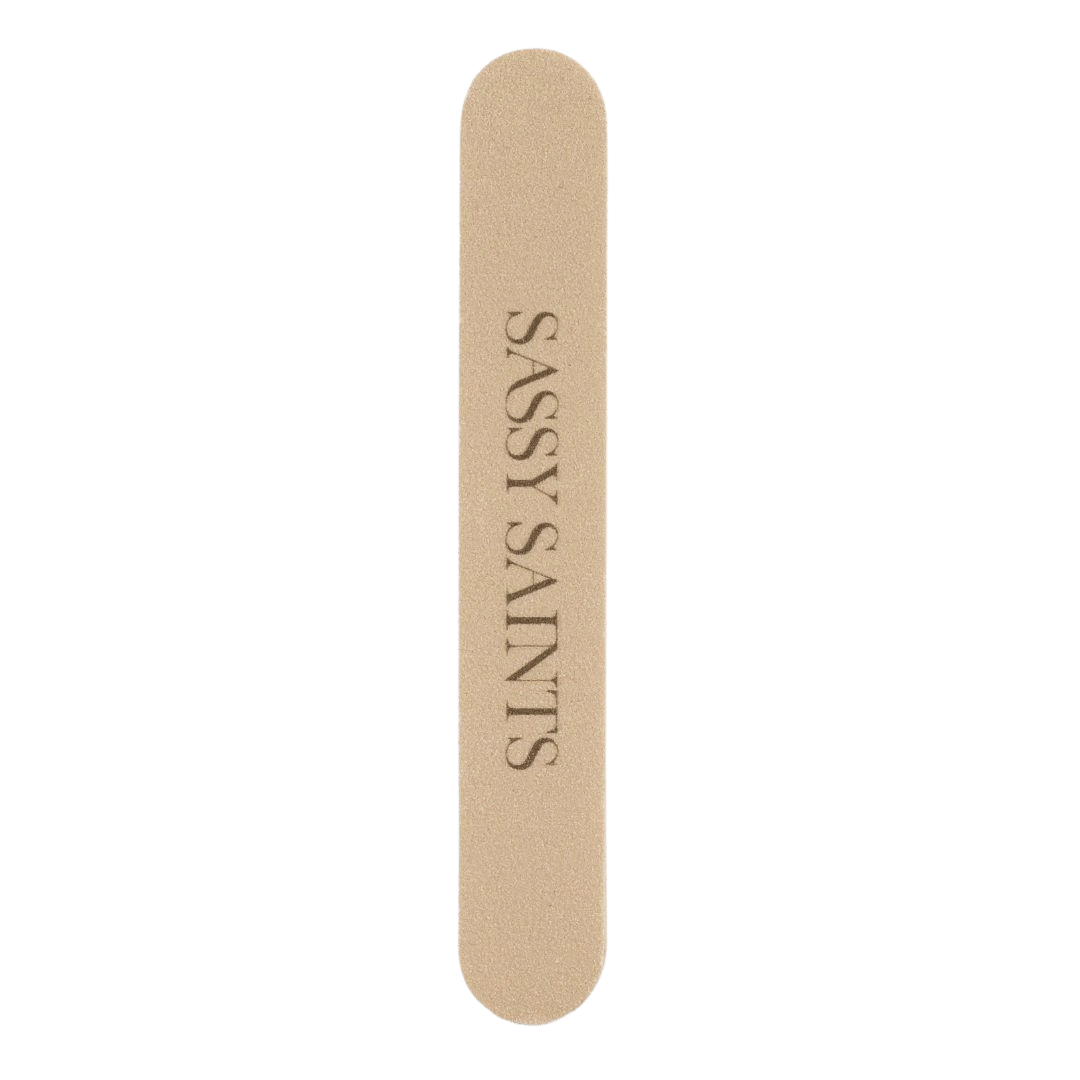 The Sassy Saints - Brings The Salon Home Nail File (FREE) is a beige, dual-grit manicure tool with rounded ends and "SASSY SAINTS" printed in bold black capital letters on its surface, making it essential for achieving flawless nails.