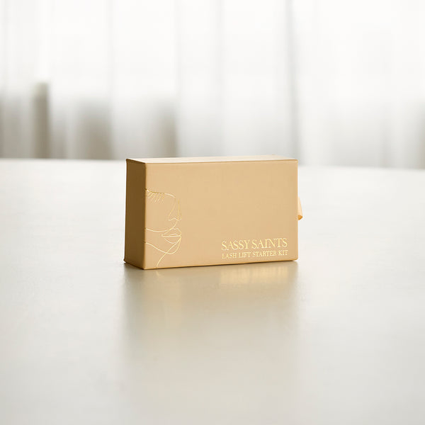 A beige rectangular box labeled Sassy Saints - Brings The Salon Home Lash Lift Mini Kit rests on a light surface with a simple face sketch; sheer curtains in the background—ideal for salon-quality curls and lifted lashes at home.
