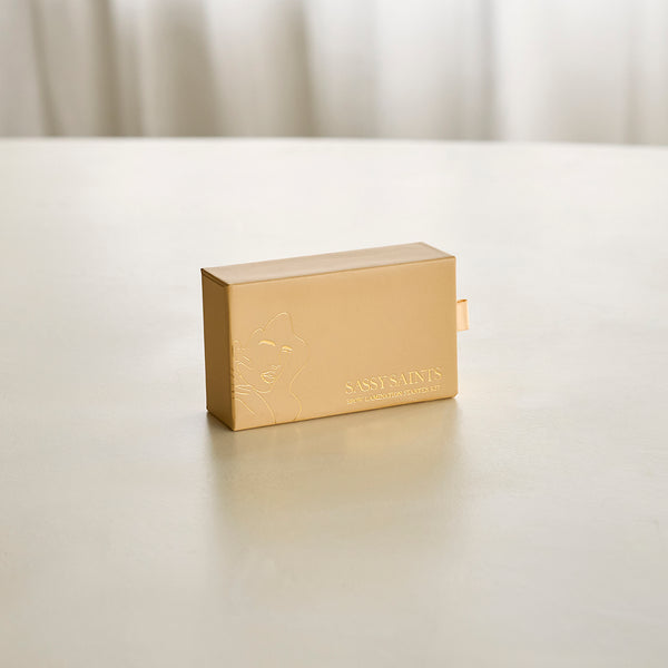 A small, gold rectangular box with subtle embossed artwork shows the text Brow Lamination Mini Kit by Sassy Saints – Brings The Salon Home, perfect for an at-home brow lamination experience, set on a smooth light surface with a neutral background.
