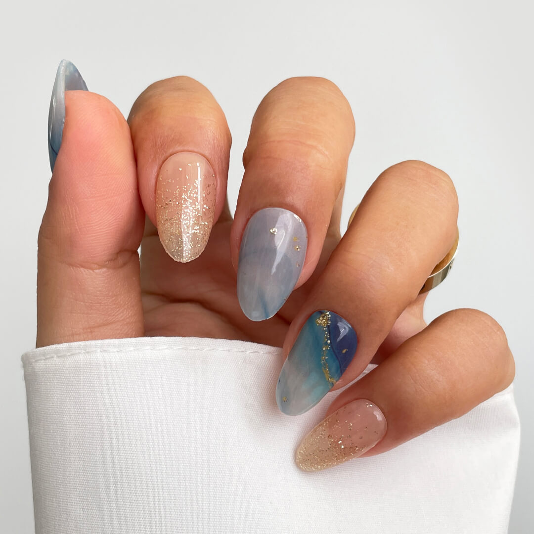 A hand with long, almond-shaped nails showcases Sassy Saints - Brings The Salon Homes Ocean Dream Gel Wrap: a creative blend of blue marble, gold glitter, and nude. Gold rings and a white shirt add elegance to this salon-quality look.