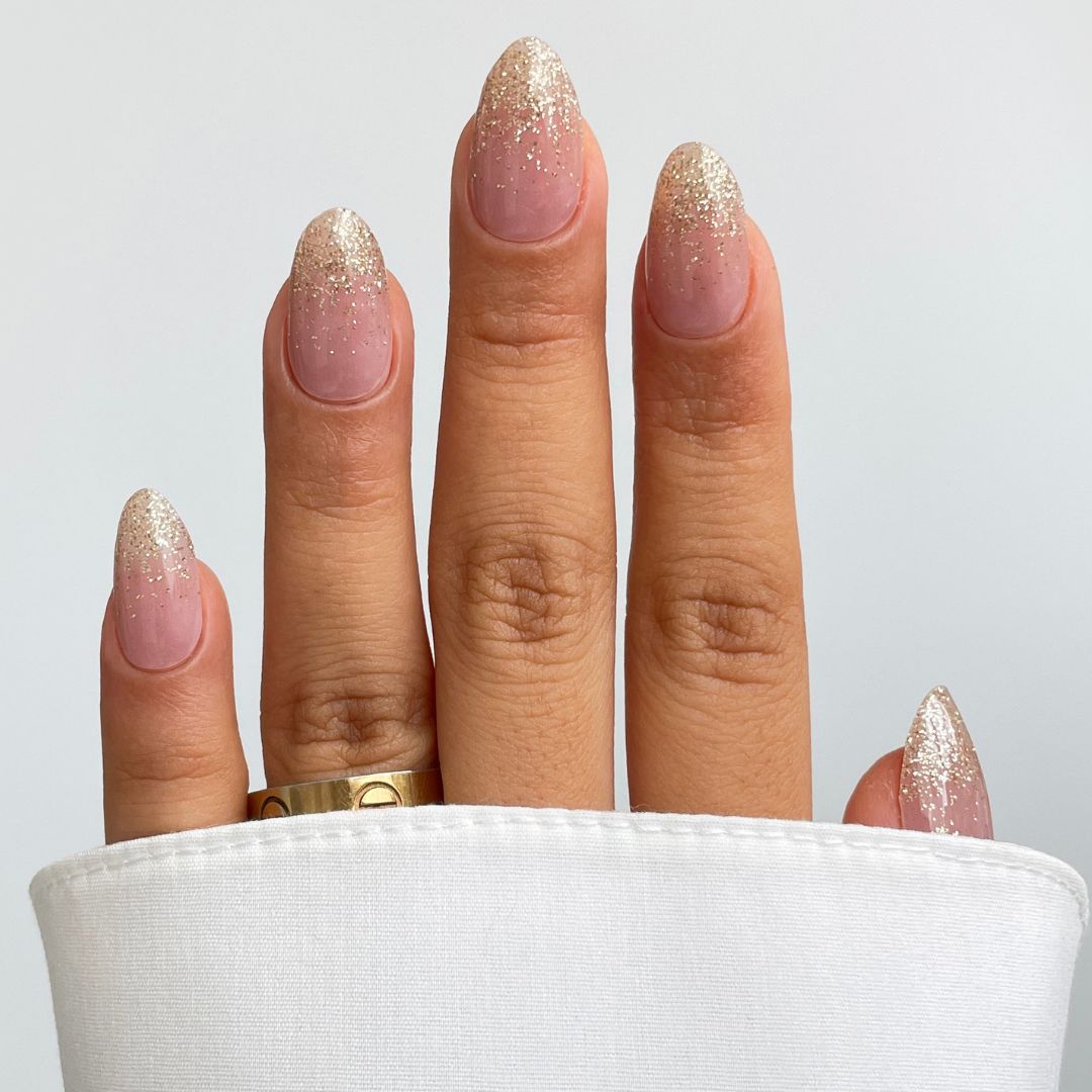 A hand shows off Sassy Saints’ Dipped In Gold Gel Wrap nails—almond-shaped with a nude base and silver glitter tips—styled with a gold ring and a white cuffed shirt against a simple background.