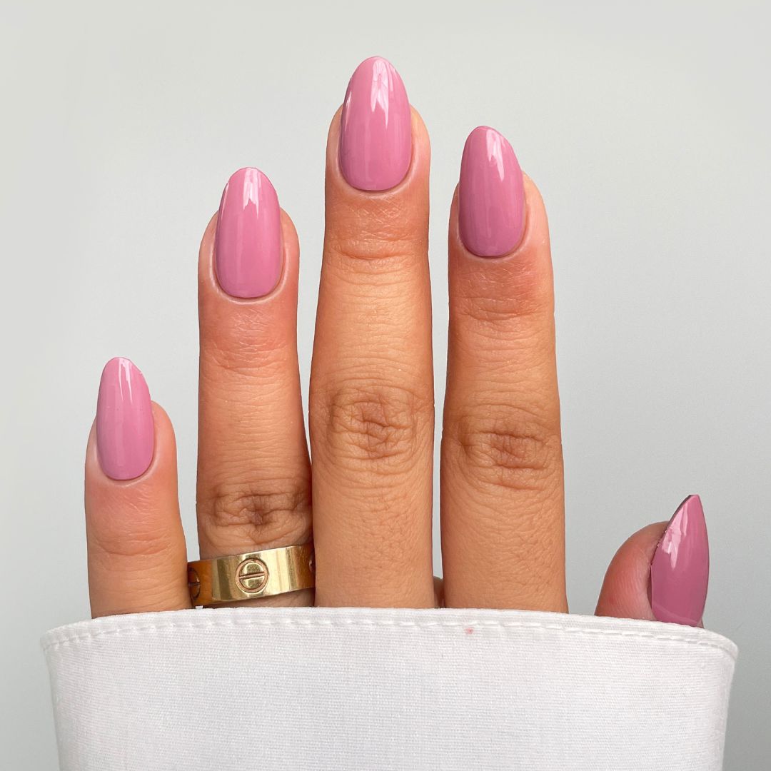 A hand with medium-length, almond-shaped nails in Sassy Saints' Dusty Blush Gel Wrap shines glossy pink. A gold ring adorns the middle finger, partially covered by a white sleeve, against a light gray background.