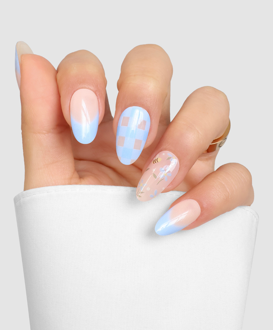 Easter Picnic by Sassy Saints - Brings The Salon Home features almond-shaped nails with pastel blue tips, a blue and white checkerboard pattern, and a floral bee design on a clear base, partially wrapped in a white cloth sleeve.