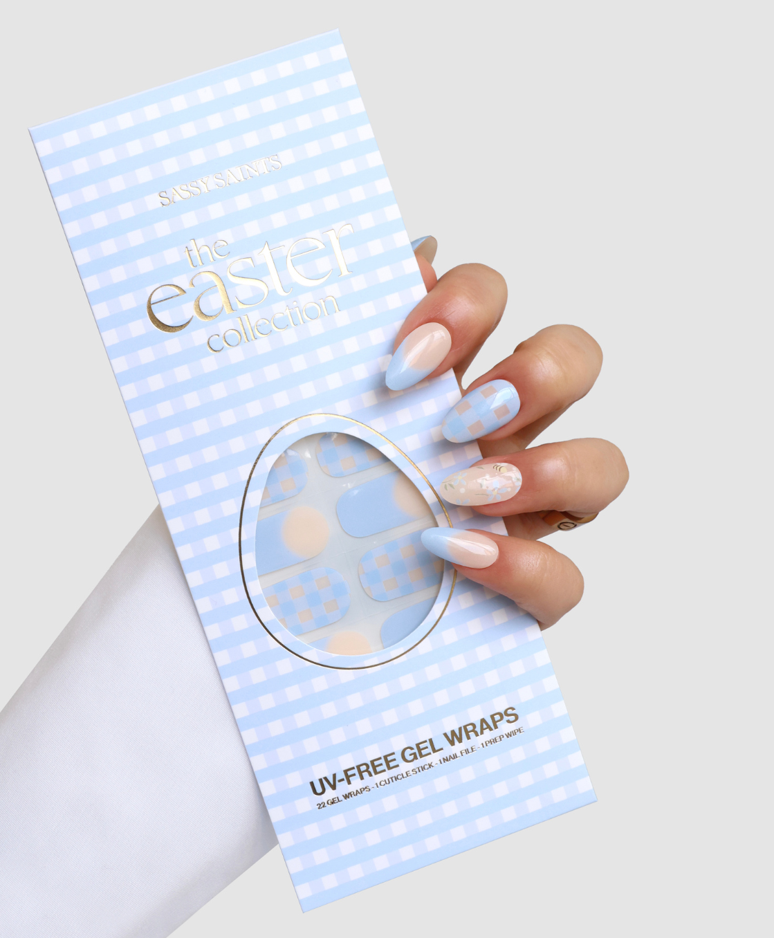 A hand with pastel blue, white, and beige patterned nails holds a Sassy Saints - Brings The Salon Home "Easter Picnic" package, featuring a blue and white gingham design with an egg-shaped window showing the UV-free gel nail wraps inside.