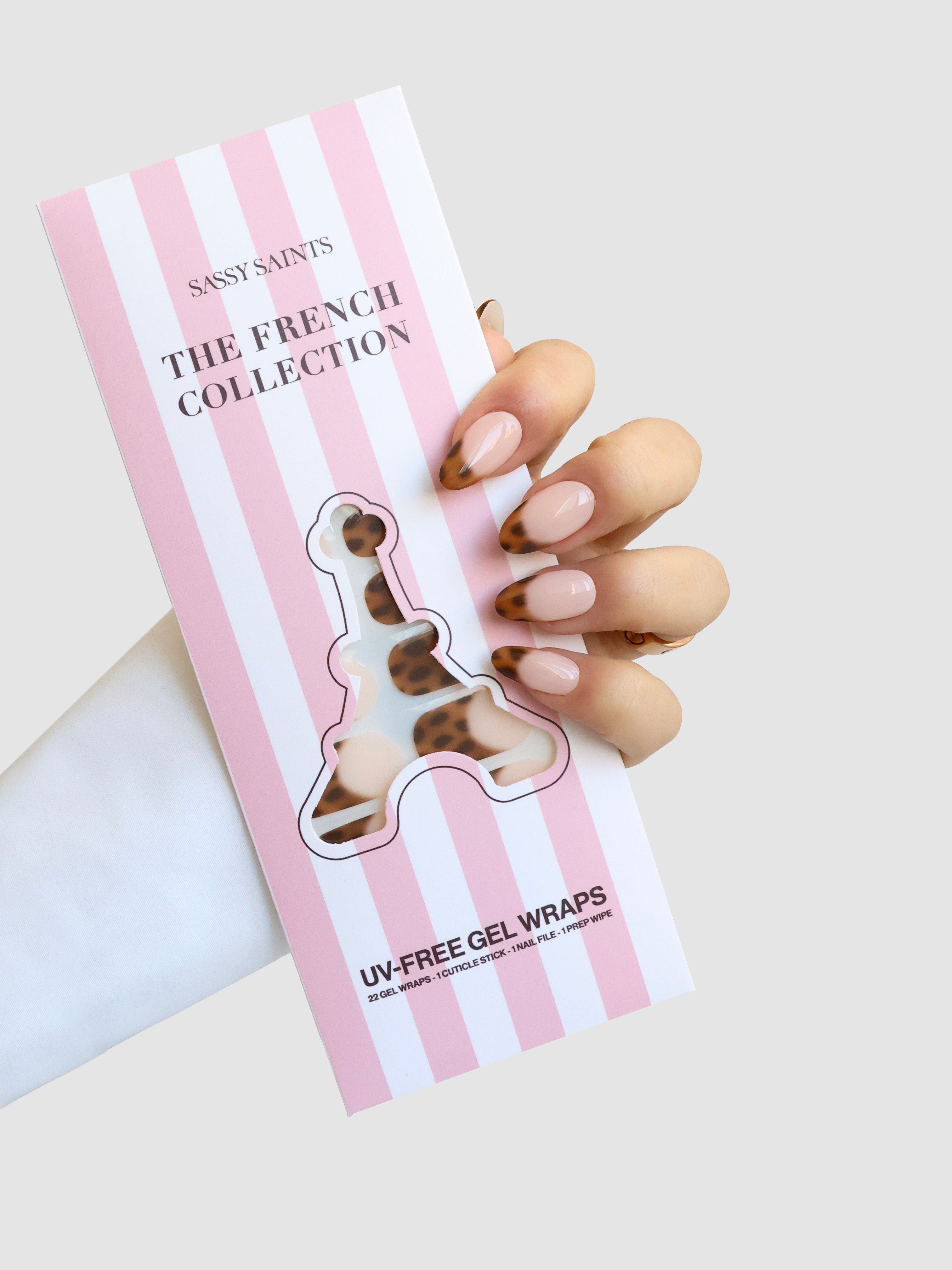 A hand with glossy nude and brown leopard-print French tips holds a pink and white striped package labeled Sassy Saints - Brings The Salon Home Écaille de Luxe Gel Wrap.
