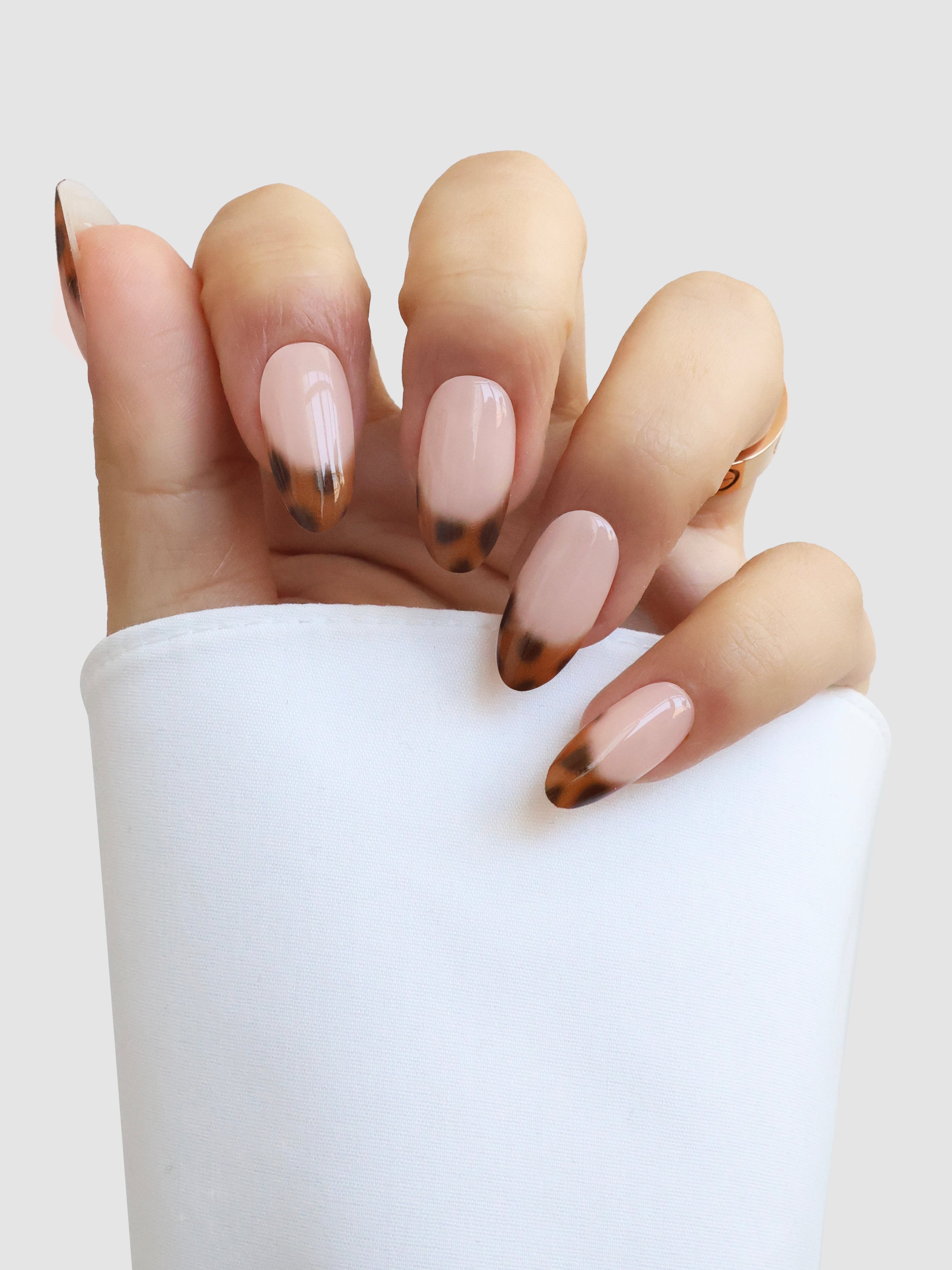 A hand showcases Sassy Saints' Écaille de Luxe Gel Wrap—almond-shaped nails with a nude base and brown tortoiseshell tips—partially wrapped in a white sleeve against a light gray background.