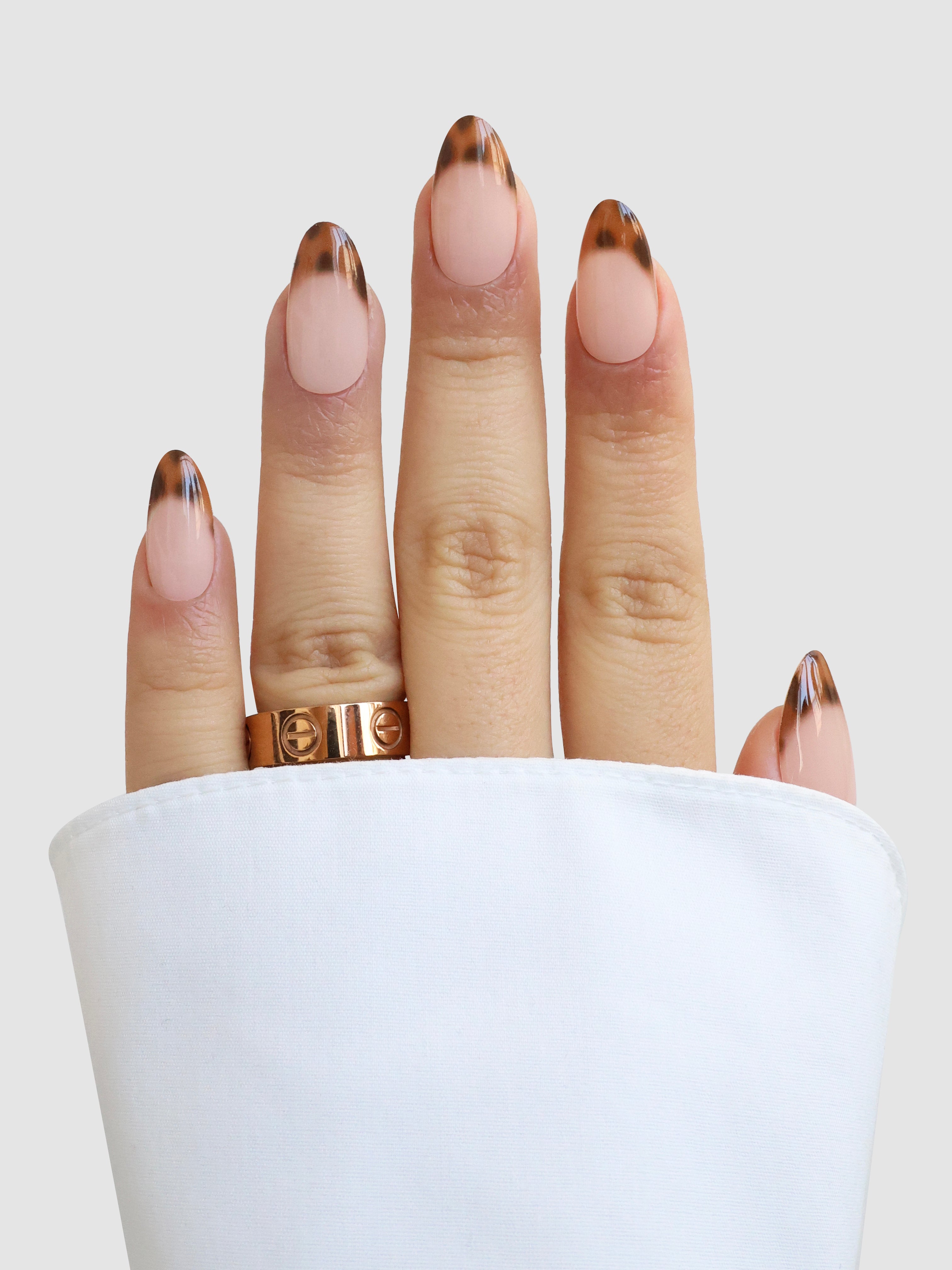 A hand with oval nails wears Écaille de Luxe Gel Wraps by Sassy Saints - Brings The Salon Home, showcasing a brown and nude abstract French tip. A gold ring and white sleeve are visible against a light gray background.