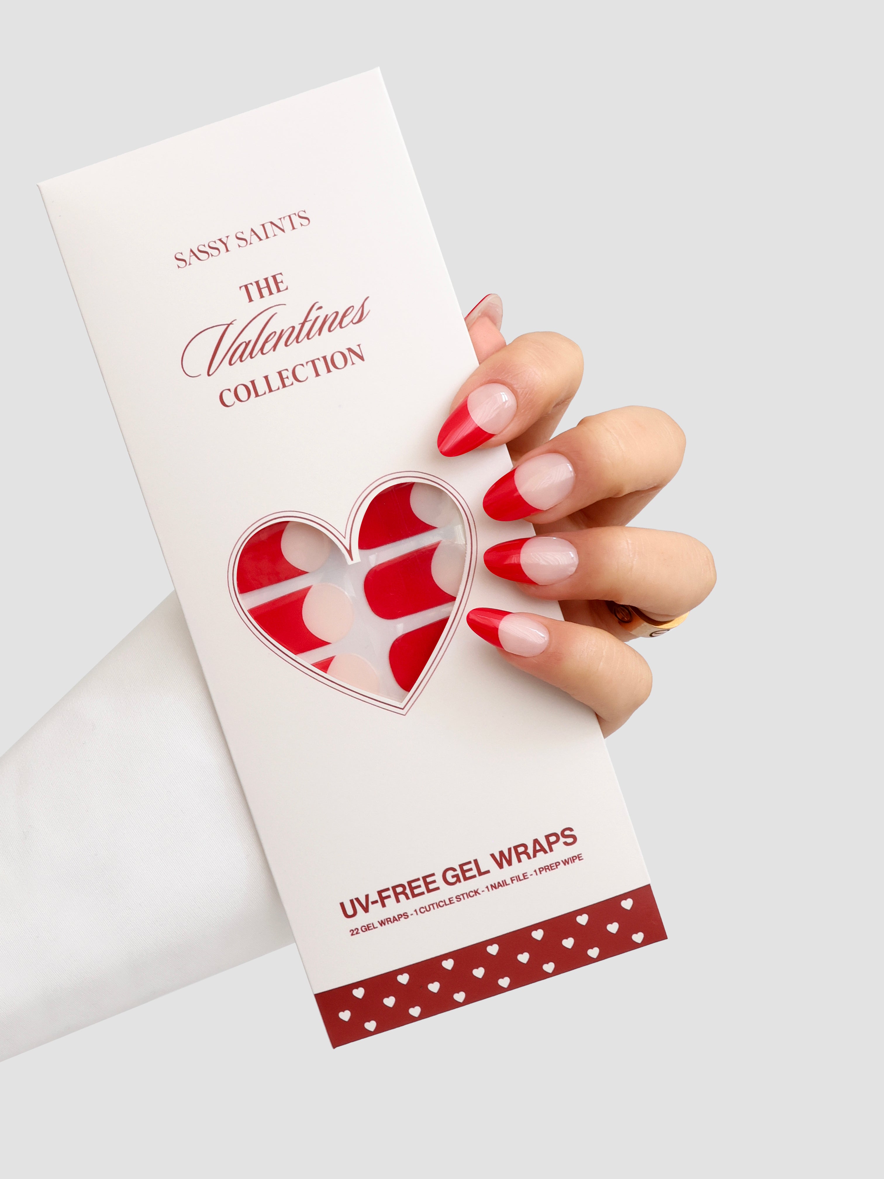 A hand with red and nude gradient nails holds a Sassy Saints - Brings The Salon Home French Kiss Gel Wrap package, with heart-shaped cutouts revealing the UV-free nail wraps inside.