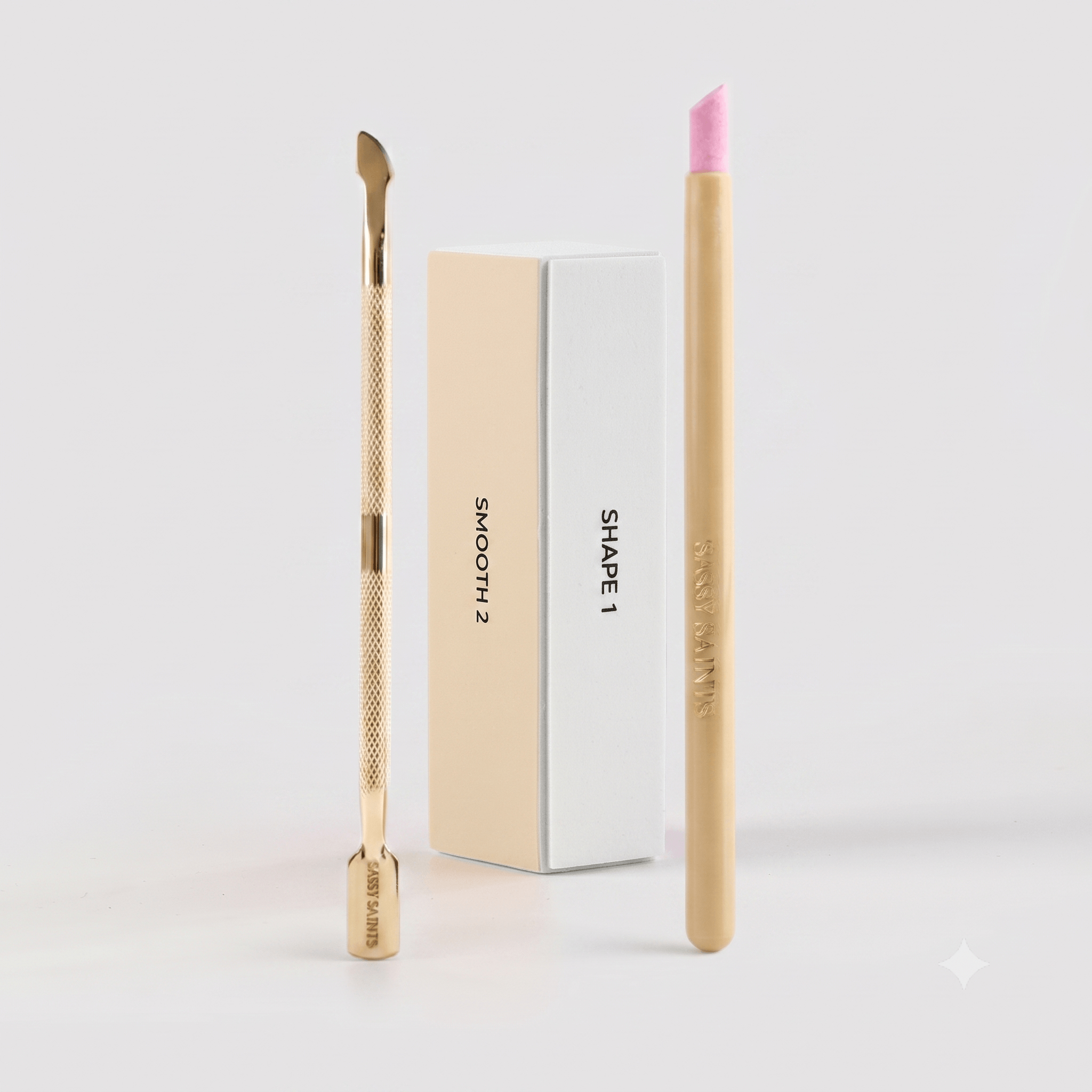 The Sassy Saints Perfect Prep Bundle—featuring a metal cuticle pusher, two rectangular nail buffer blocks labeled “SMOOTH 2” and “SHAPE 1,” and a beige nail file with a pink tip—is arranged against a white background.