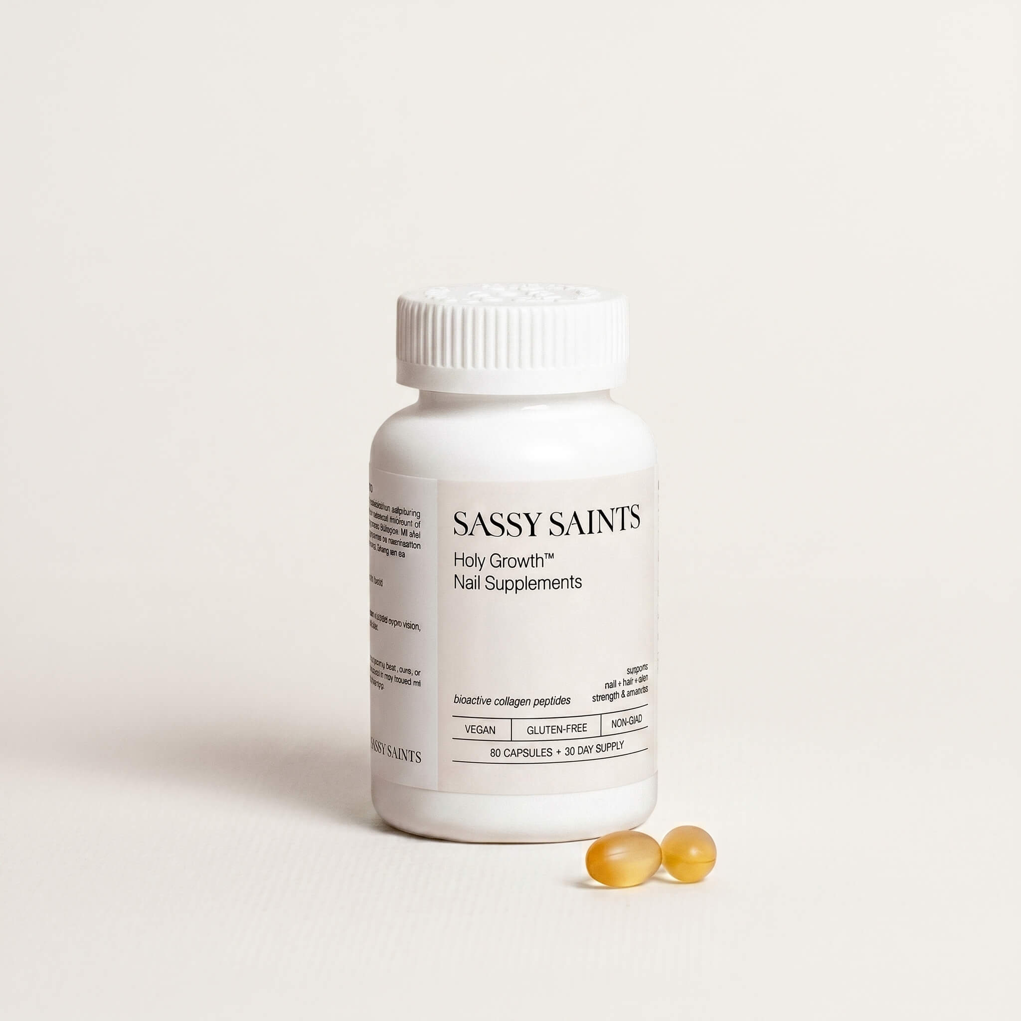 A white bottle labeled Biotin Nail Growth Supplement by Sassy Saints - Brings The Salon Home stands upright on a light background, with two yellow capsules placed beside it.