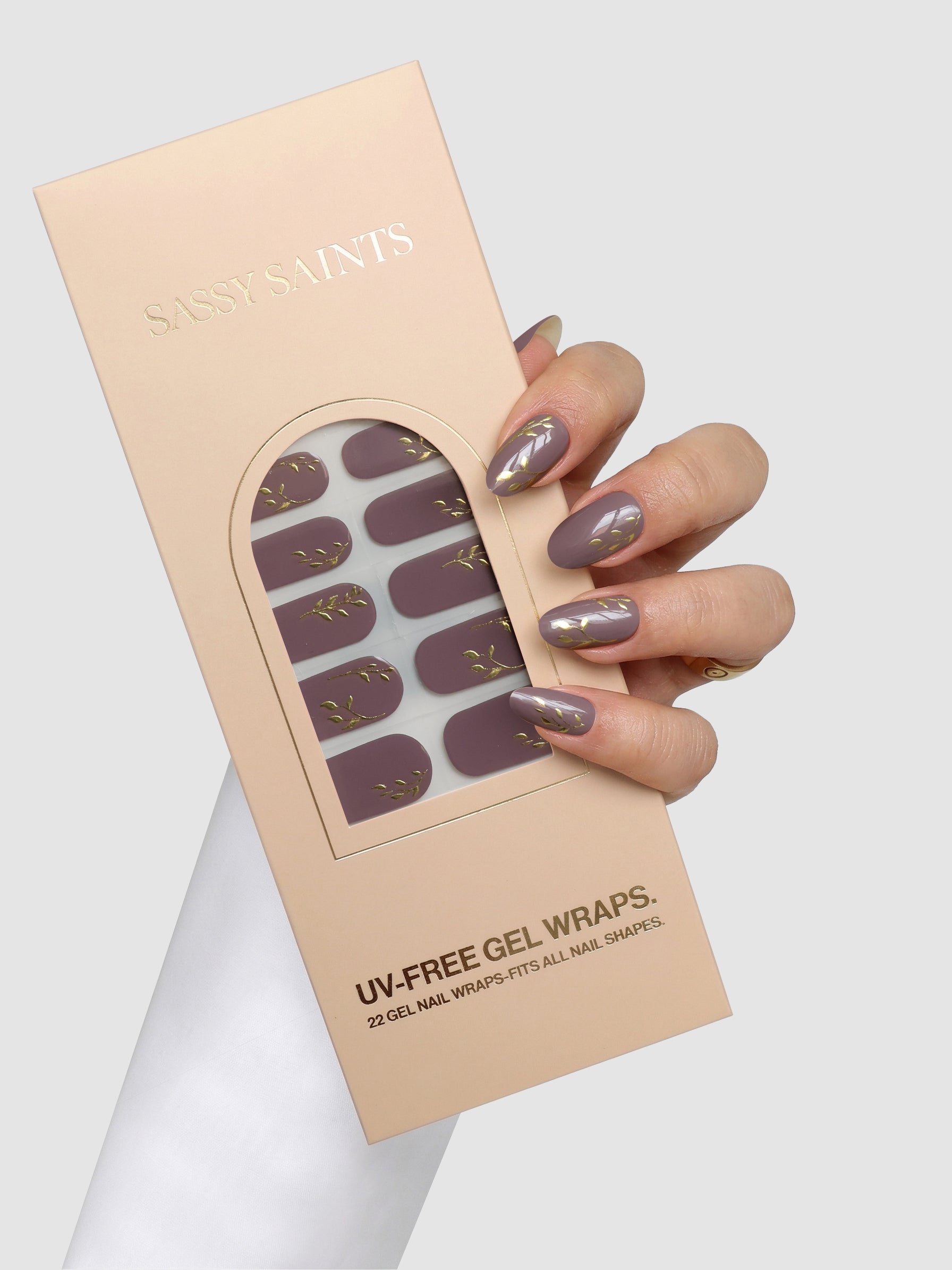 A hand with mauve and gold-accented nails holds a box of Sassy Saints - Brings The Salon Home Gilded Mauve Gel Wraps. The light pink box features a window displaying the mauve wraps adorned with elegant gold details.