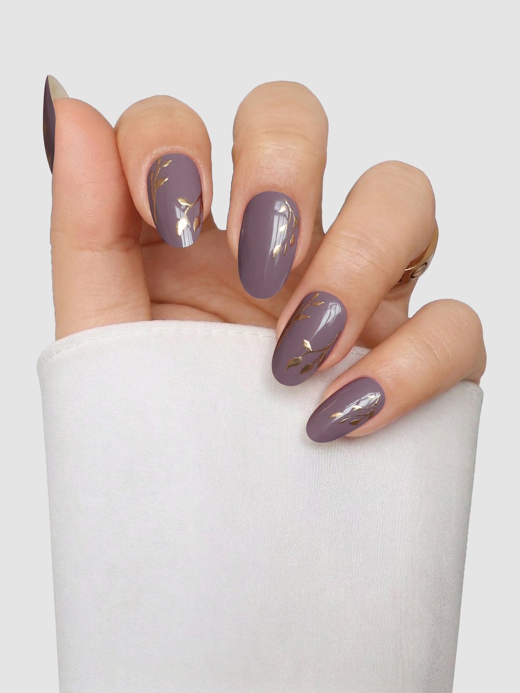 A hand displays Gilded Mauve Gel Wrap nail covers by Sassy Saints - Brings The Salon Home, shaped into an elegant almond style and featuring mauve tones with delicate gold leaf accents on two nails. The background is plain and light, with the wrist wrapped in a white fabric.