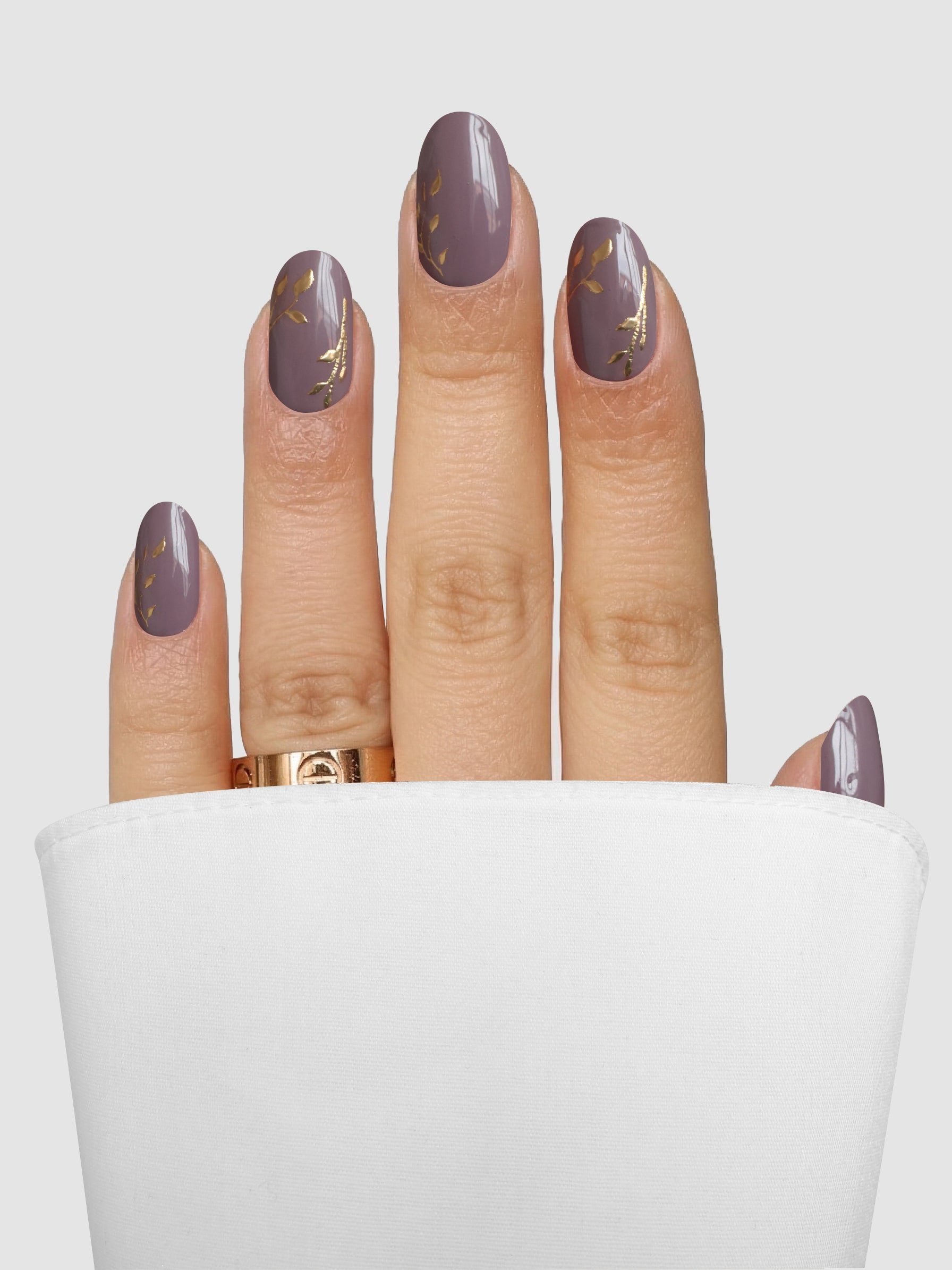 A hand showcases Sassy Saints - Brings The Salon Home’s Gilded Mauve Gel Wrap nails with elegant gold leaf designs and a matching gold ring, partially wrapped in white fabric against a light gray background.