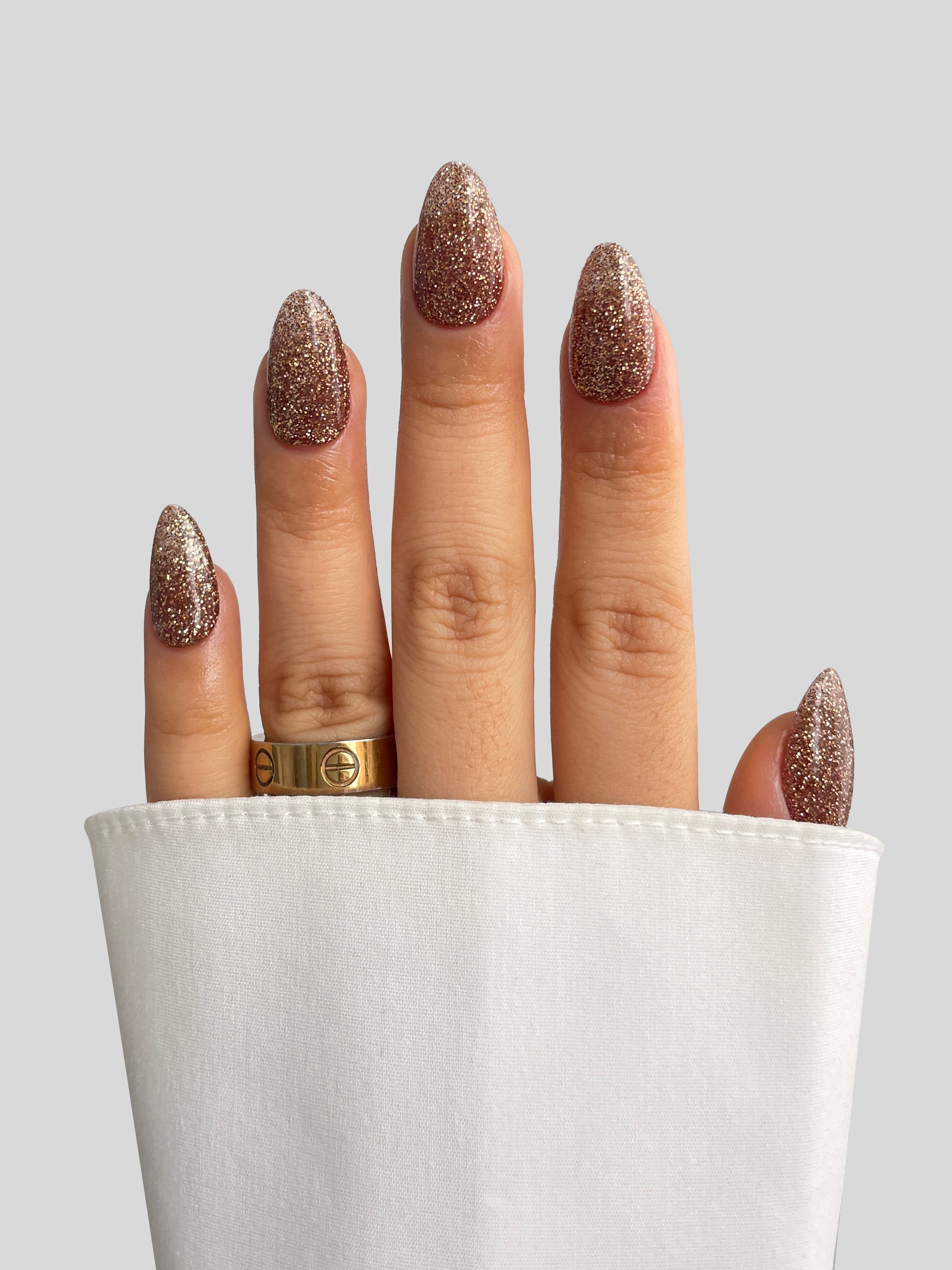 A hand with long, glittery gold nails created using Sassy Saints' Golden Gift Dipping Powder and adorned with a gold ring is partially wrapped in a white cloth, set against a plain light gray background.
