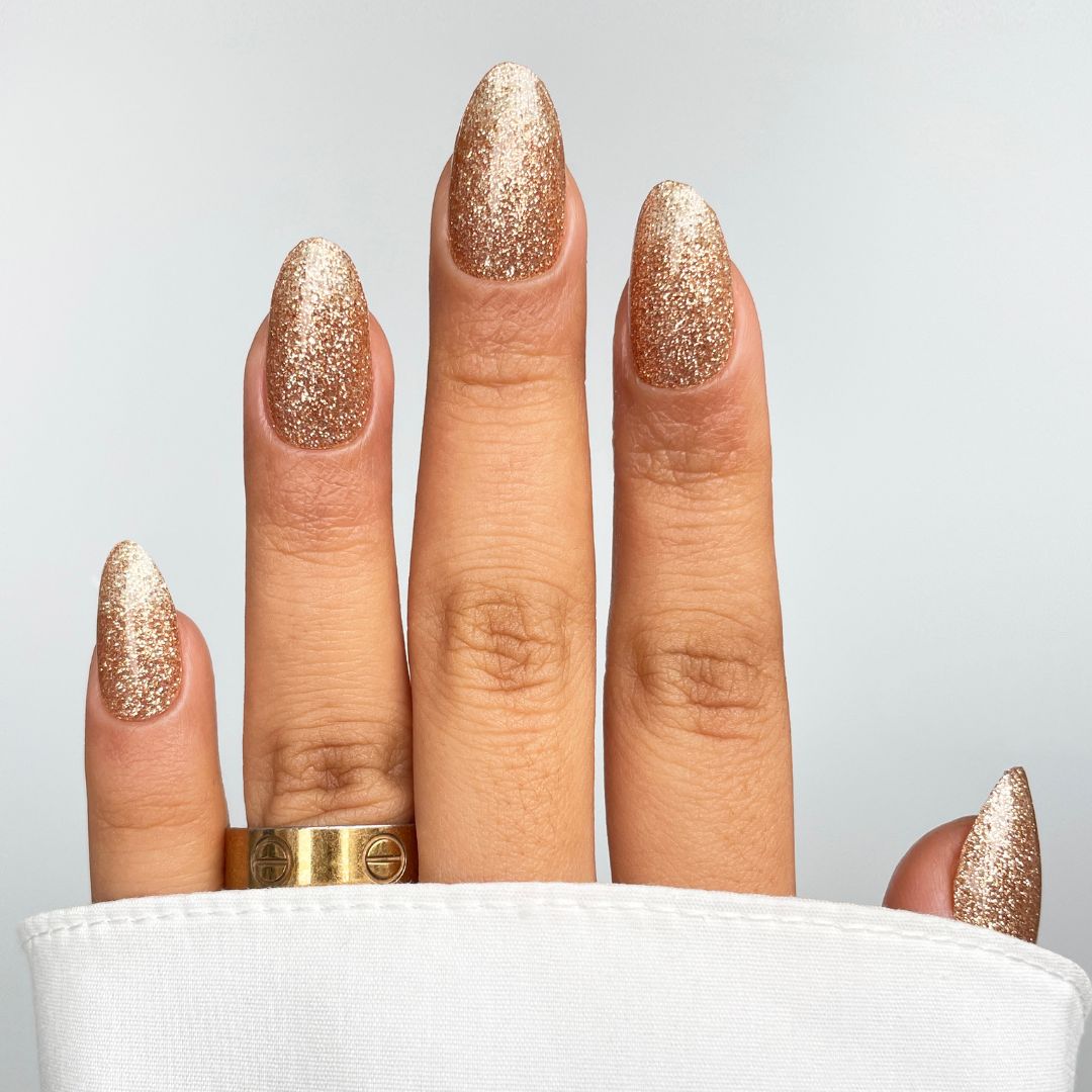 A hand with almond-shaped nails in Sassy Saints - Brings The Salon Home Golden Hour Gel Wrap, wearing a gold ring on the middle finger, is partially covered by white fabric against a plain light gray background.