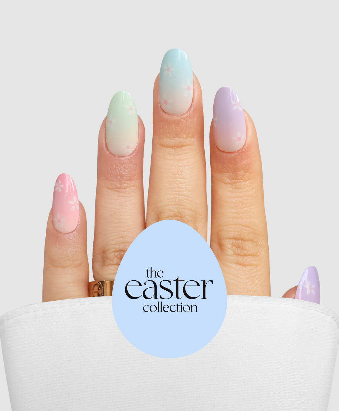 A hand with pastel almond-shaped nails from Sassy Saints - Brings The Salon Home’s Blooming Days, adorned with white floral art and partly veiled by white cloth. “The Easter Collection” appears in text on a blue egg shape.