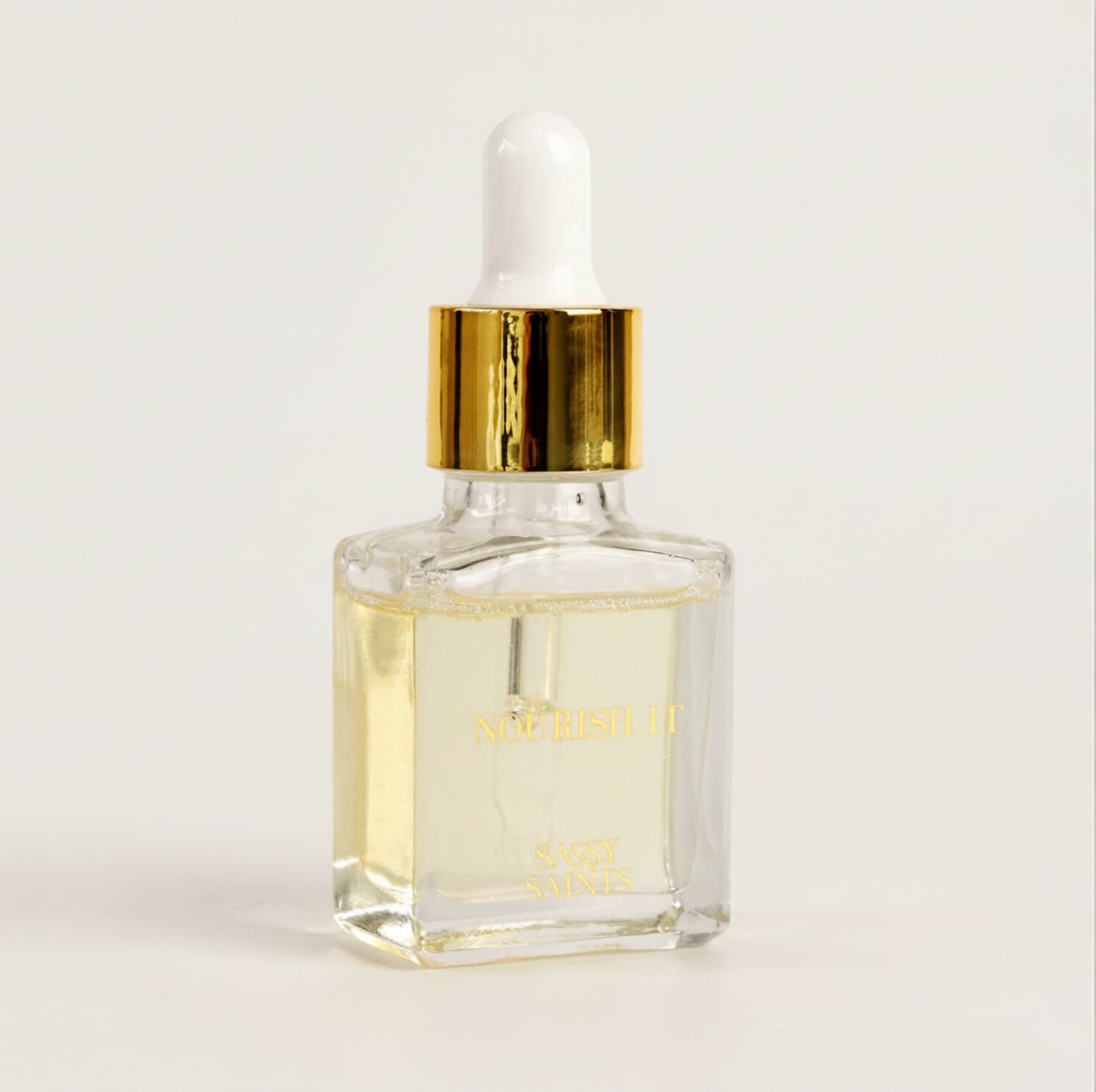 A clear glass dropper bottle with a gold and white cap holds light yellow Cuticle Oil from Sassy Saints – Brings The Salon Home. The label says "Nourish It" in gold, perfect for nail growth and healthy cuticles.