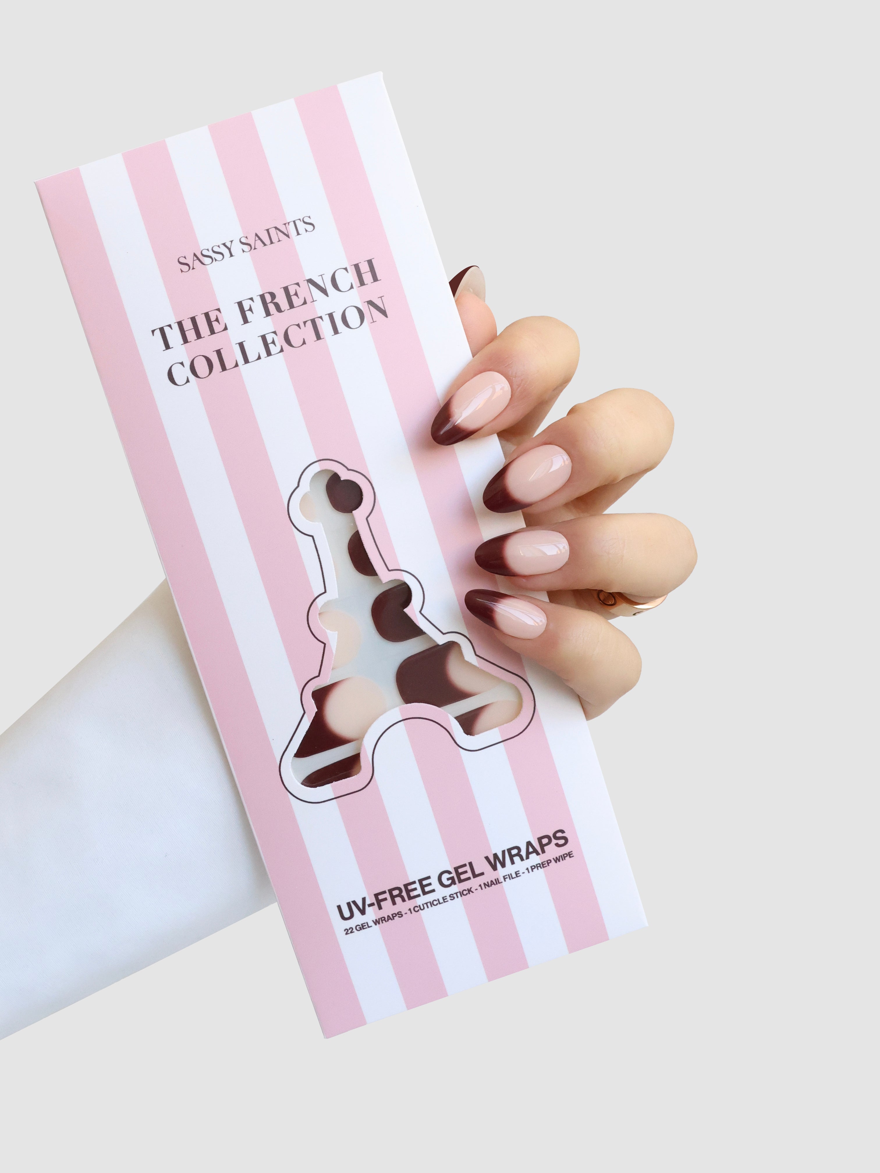 A hand with pink nude manicured nails holds a box labeled Sassy Saints - Brings The Salon Home Haute Bordeaux Gel Wrap, featuring a cutout displaying the nail wrap colors inside.