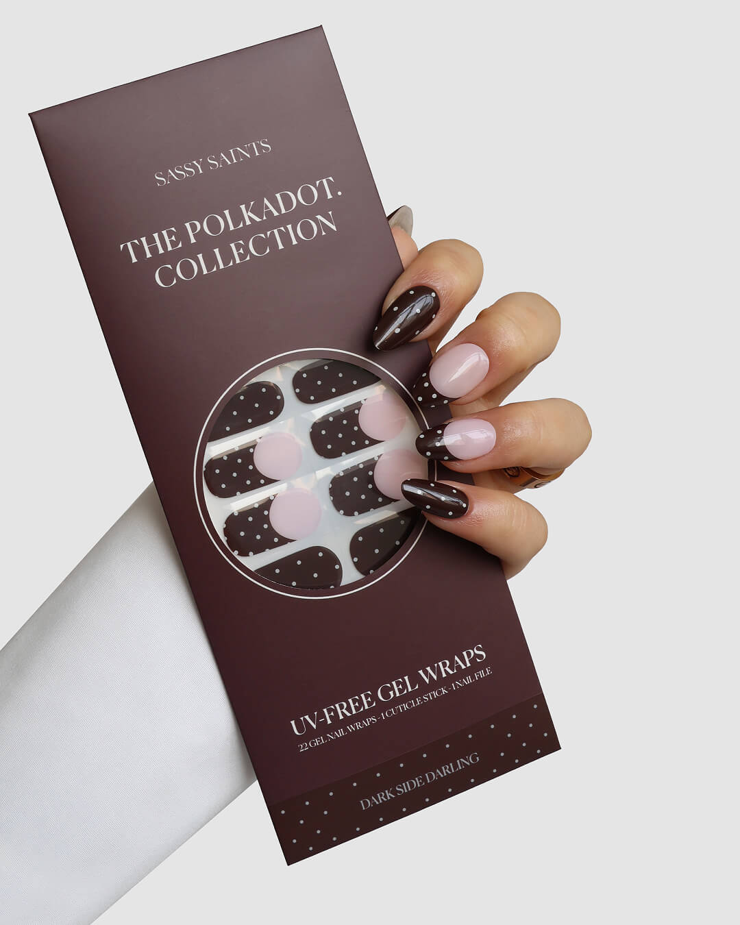 A hand with dark brown and pale pink manicured nails holds a maroon Sassy Saints - Brings The Salon Home package labeled Dark Side Darling Gel Wrap, featuring polka dot UV-free gel nail wraps.