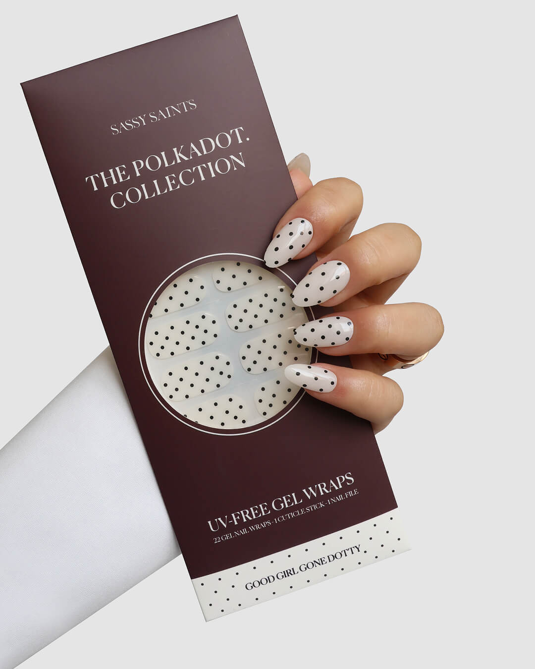 A hand with long, white nails adorned with black polka dots holds a burgundy package labeled Sassy Saints - Brings The Salon Home Good Girl Gone Dotty Gel Wrap, featuring matching UV-free nail wraps inside.