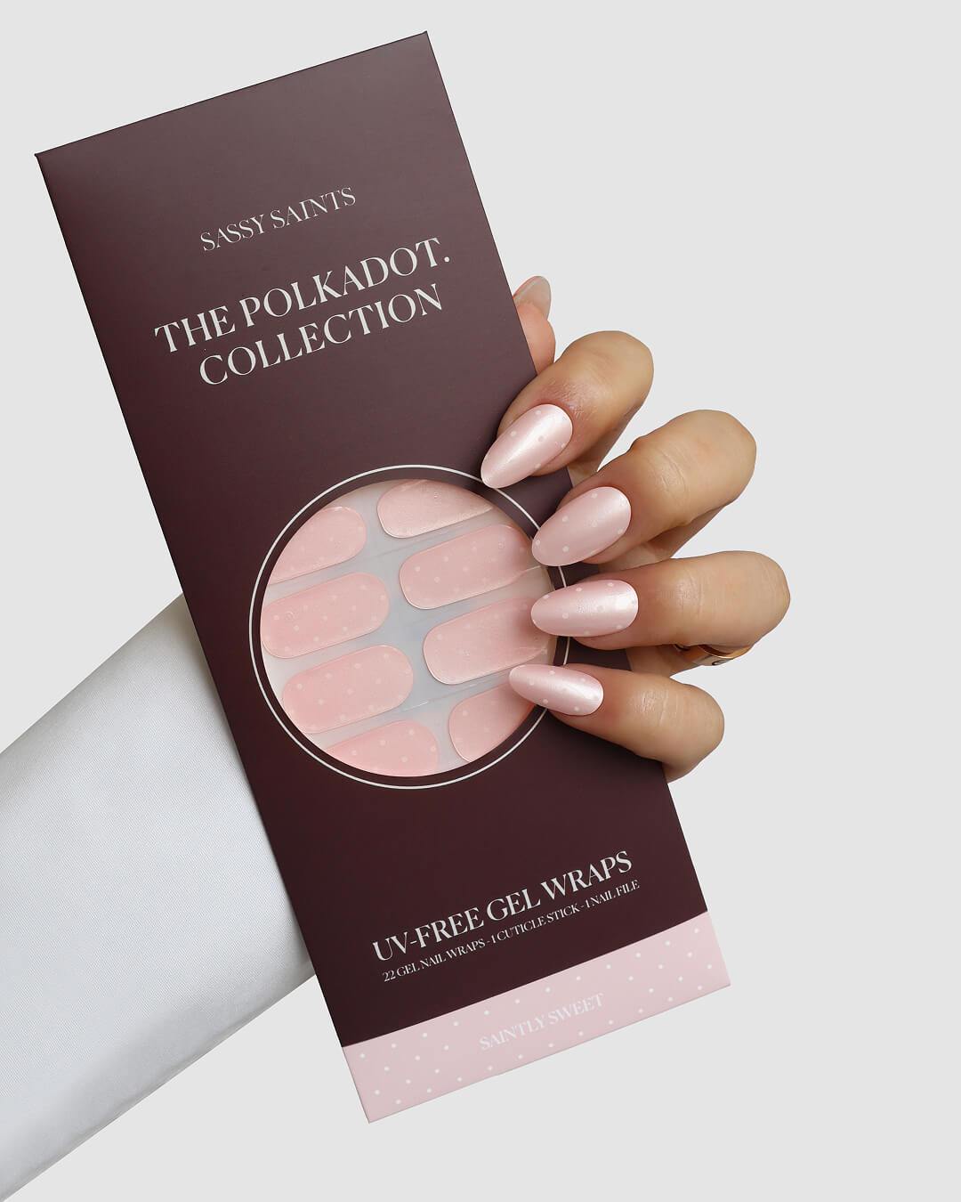 A hand with long, glossy, light pink nails holds a maroon Saintly Sweet Gel Wrap package by Sassy Saints - Brings The Salon Home, containing matching UV-free gel nail wraps with pink polka dots.