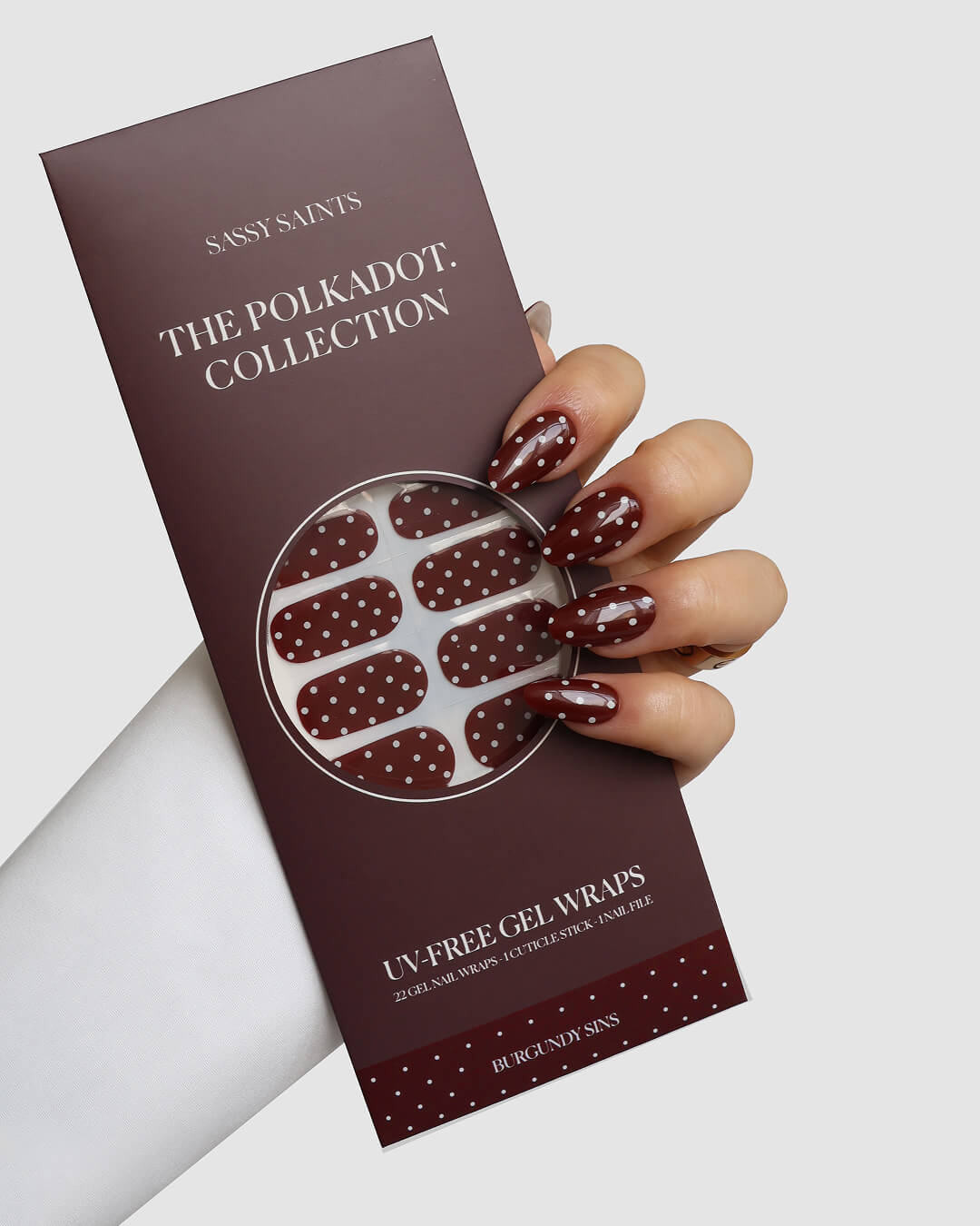 A hand with long, oval nails painted burgundy with white polka dots holds a matching pack of Sassy Saints - Brings The Salon Home’s Burgundy Sins Gel Wraps.
