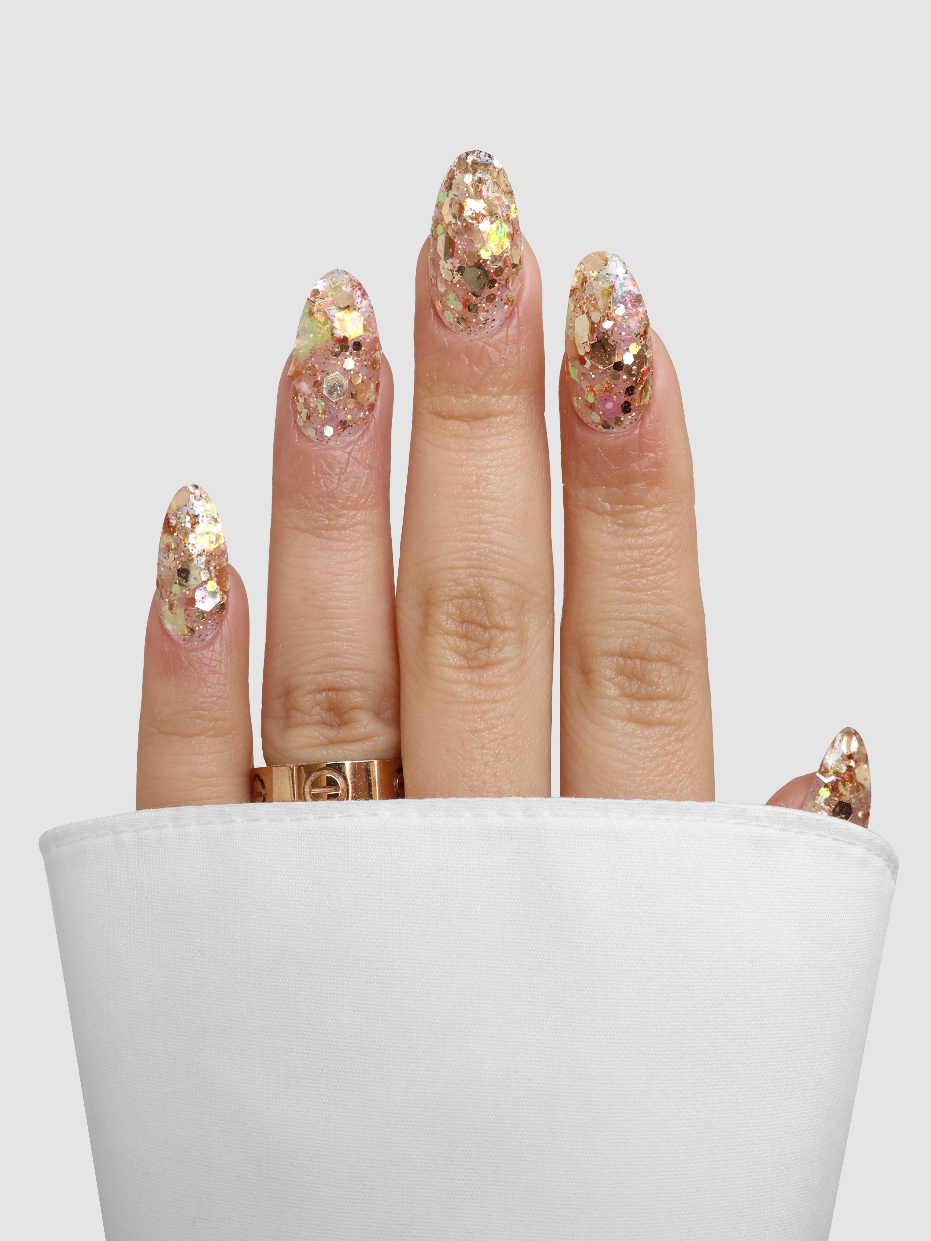 A hand with long, almond-shaped nails painted in Sassy Saints - Brings The Salon Home’s Honey Glaze Dipping Powder rests on a white surface. The person wears a gold ring and partially tucks their nails into a white cloth.