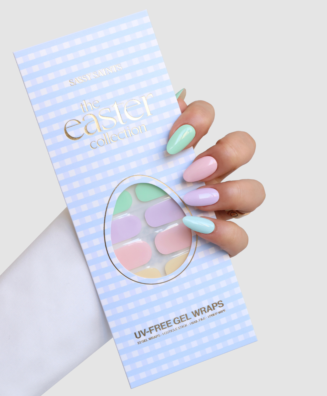 A hand with pastel nails holds a package of Sassy Saints' "Hop Into Spring" UV-free gel wraps in pastel shades, from their Easter collection, against a light gray background.