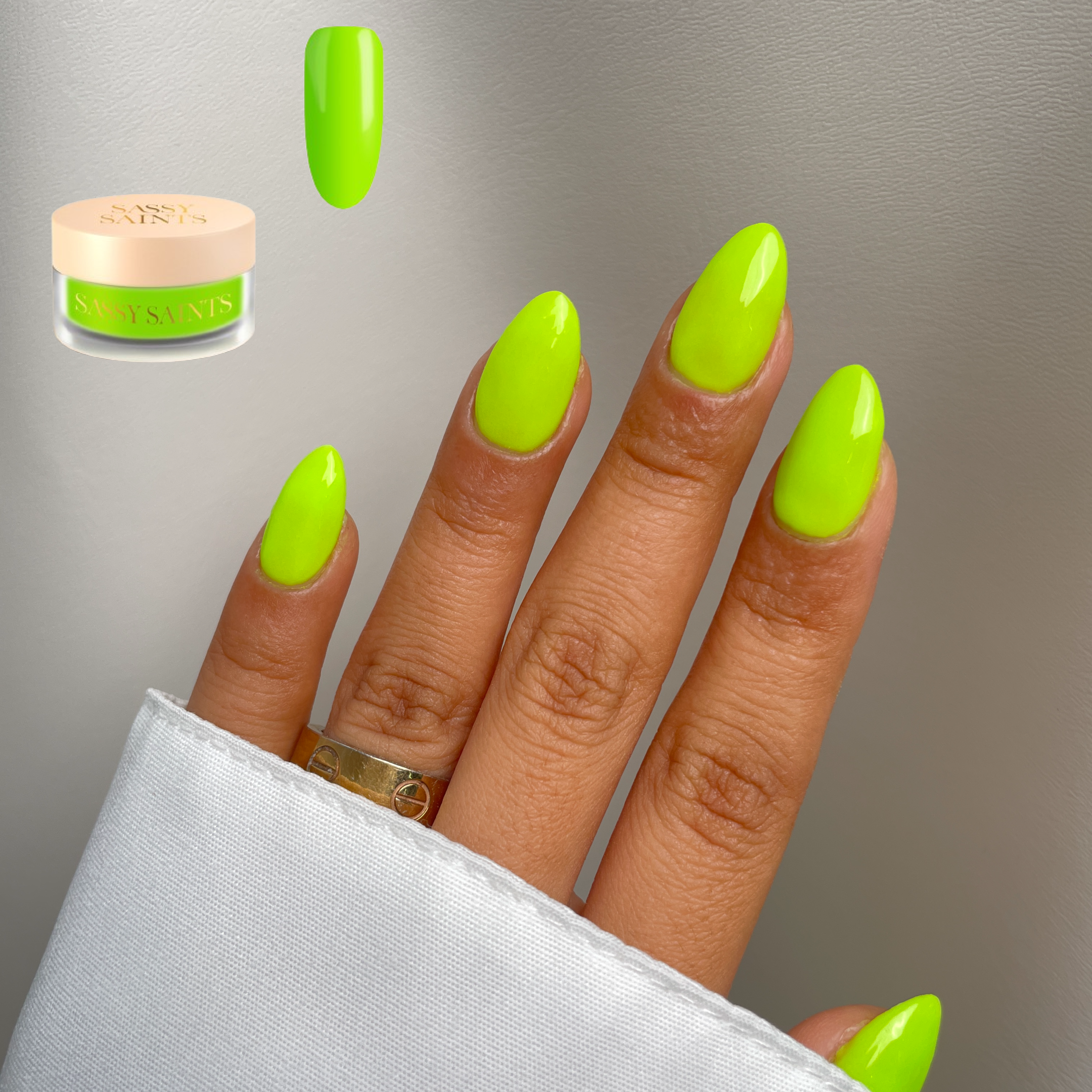 A hand with neatly manicured nails shines in Sassy Saints - Brings The Salon Homes Lime Light Dipping Powder, a vivid neon yellow-green. A gold ring accents the look, with a matching powder jar and sample nail tip displayed nearby.