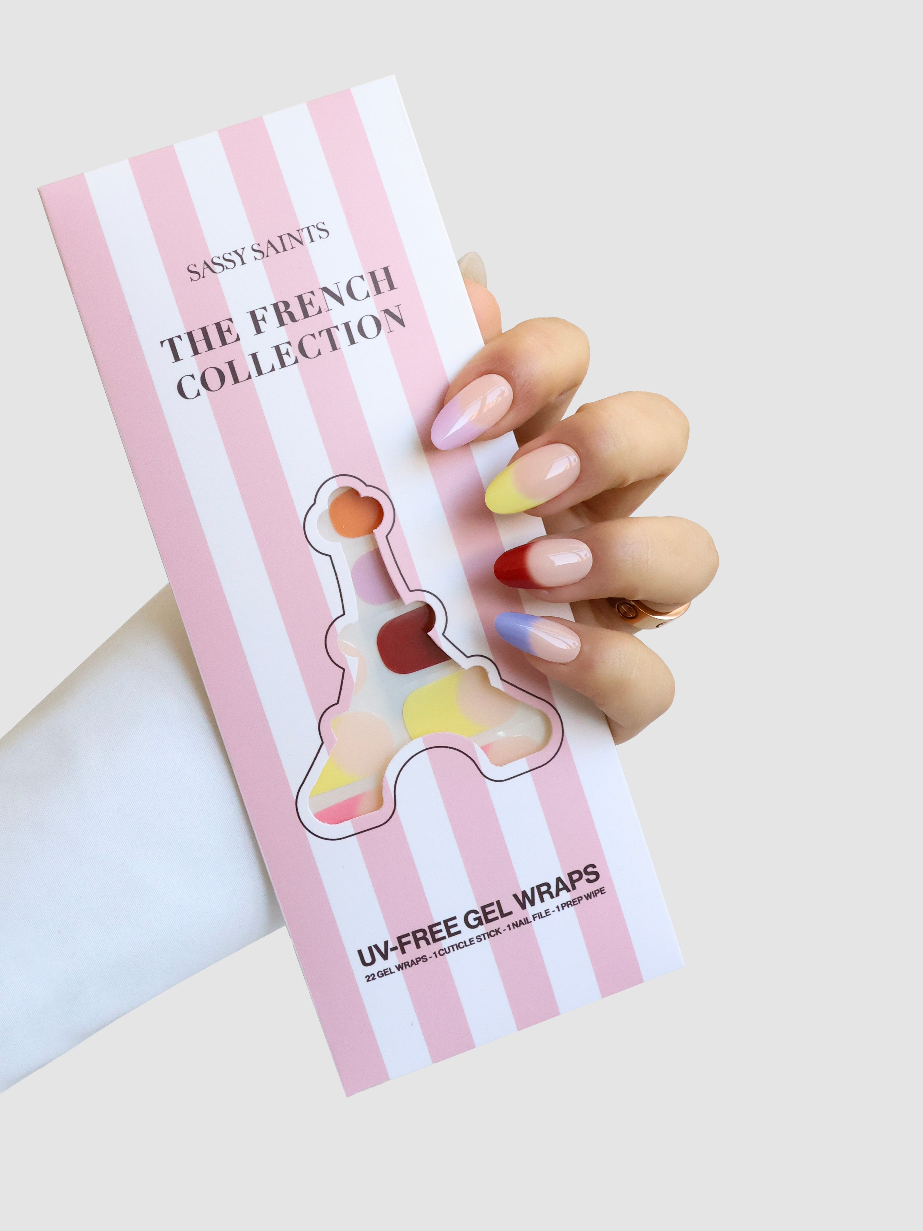 A hand with pastel pink manicured nails holds a pink and white striped package labeled Sassy Saints - Brings The Salon Home Jardin de Couleurs Gel Wrap, featuring an Eiffel Tower-shaped cutout.