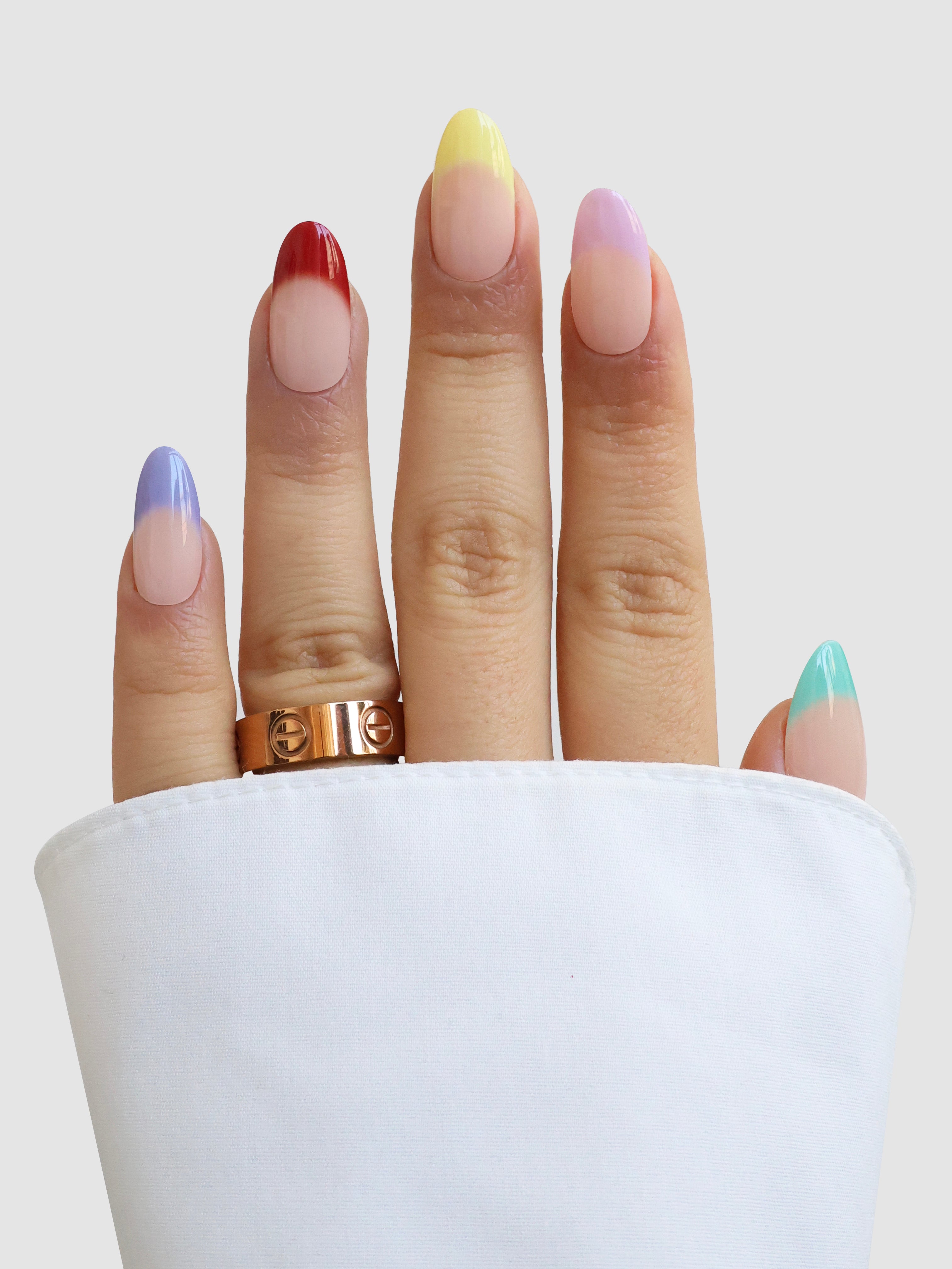 A hand with Jardin de Couleurs Gel Wrap nails from Sassy Saints - Brings The Salon Home—each nail a different ombre shade—emerges from a white sleeve, adorned with a gold ring on the middle finger.