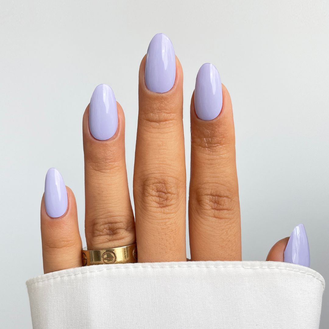 A hand with neatly manicured nails featuring Sassy Saints’ Lavender Blush Gel Wrap, a glossy pastel lavender, wears a gold ring on the ring finger and is partly covered by a white sleeve against a light background.