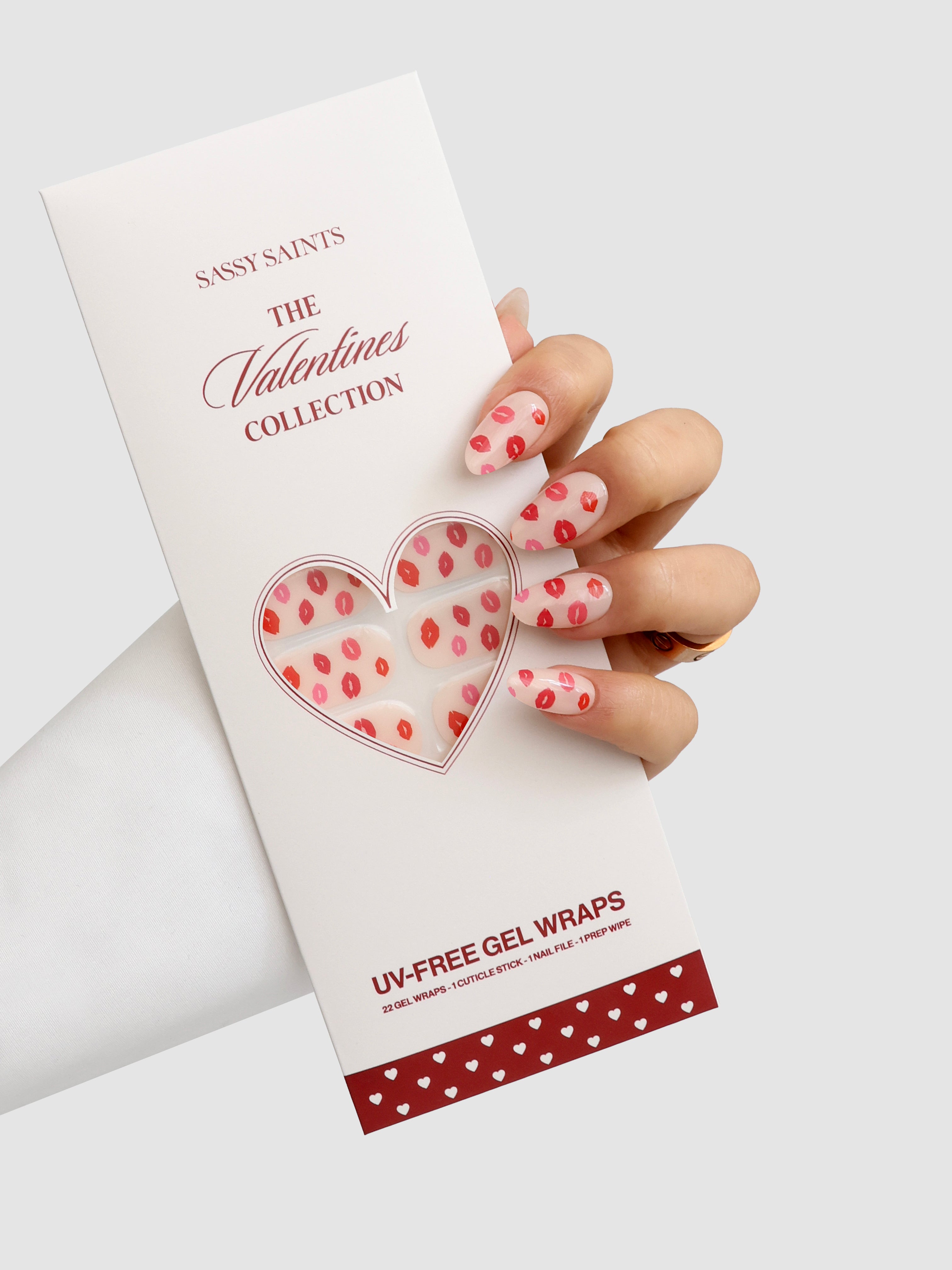 A hand with pink, lip-patterned nail wraps holds a white Sassy Saints - Brings The Salon Home package labeled Lip Service Gel Wrap, featuring a heart-shaped window displaying the matching wraps inside.