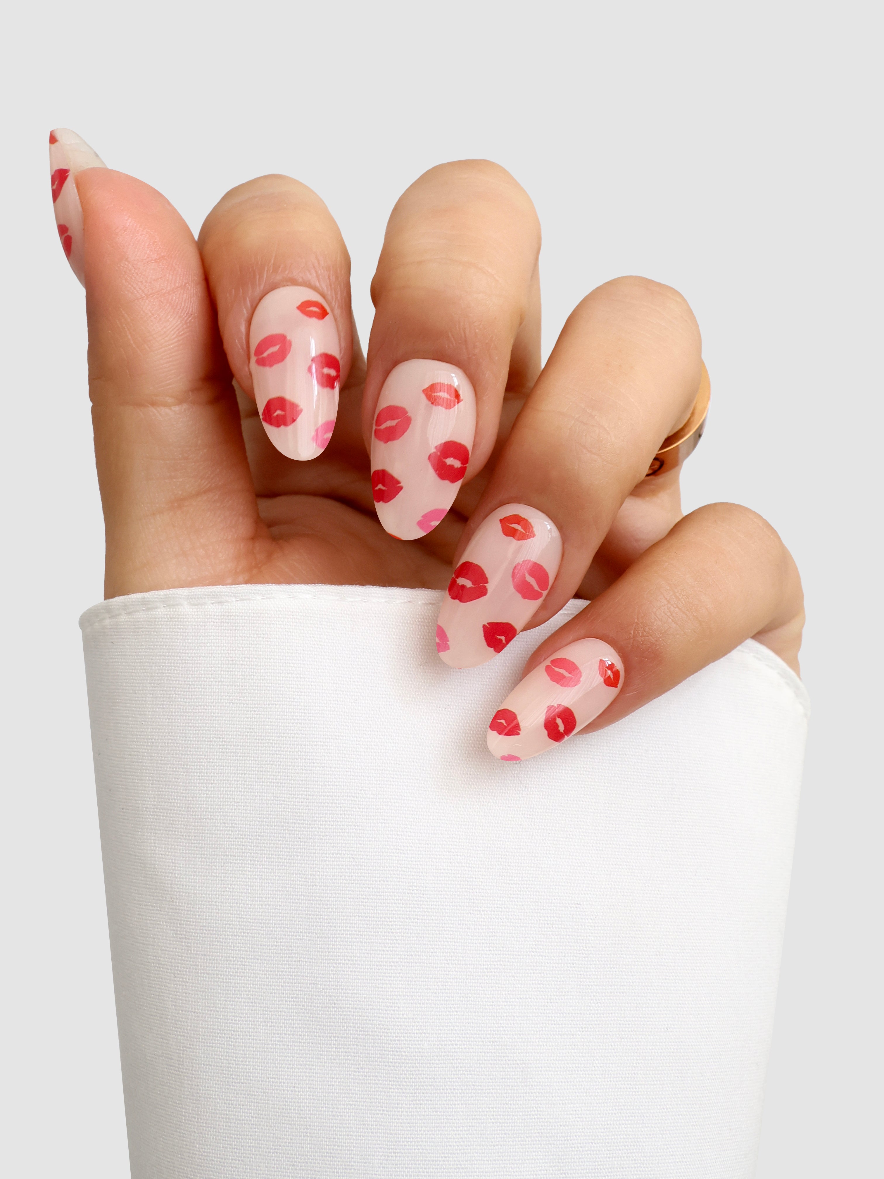 A hand features Sassy Saints’ Lip Service Gel Wrap—long, almond-shaped nails with a clear base and red lips design—partly covered by a white sleeve, set against a plain light background.