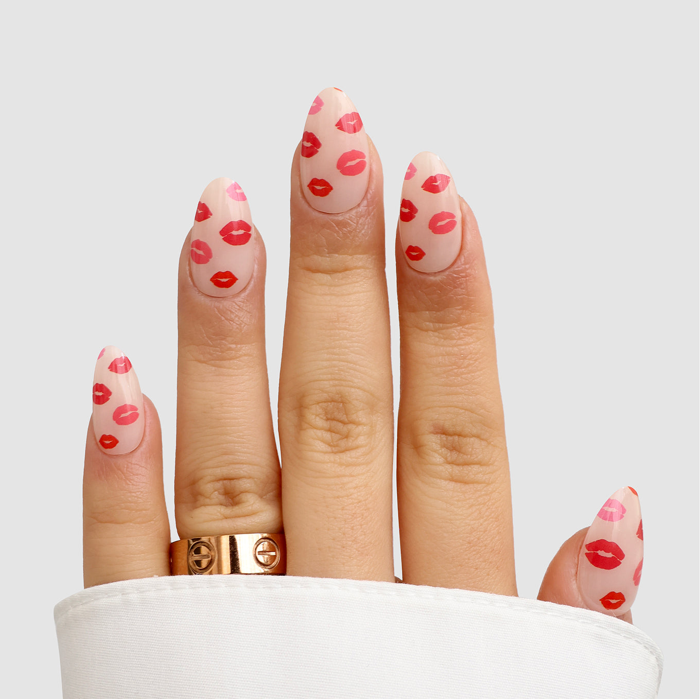 A hand shows off Sassy Saints - Brings The Salon Home Lip Service Gel Wraps: long, almond nails in pale pink with tiny red lips, paired with a gold ring and white sleeve, set against a light gray background.
