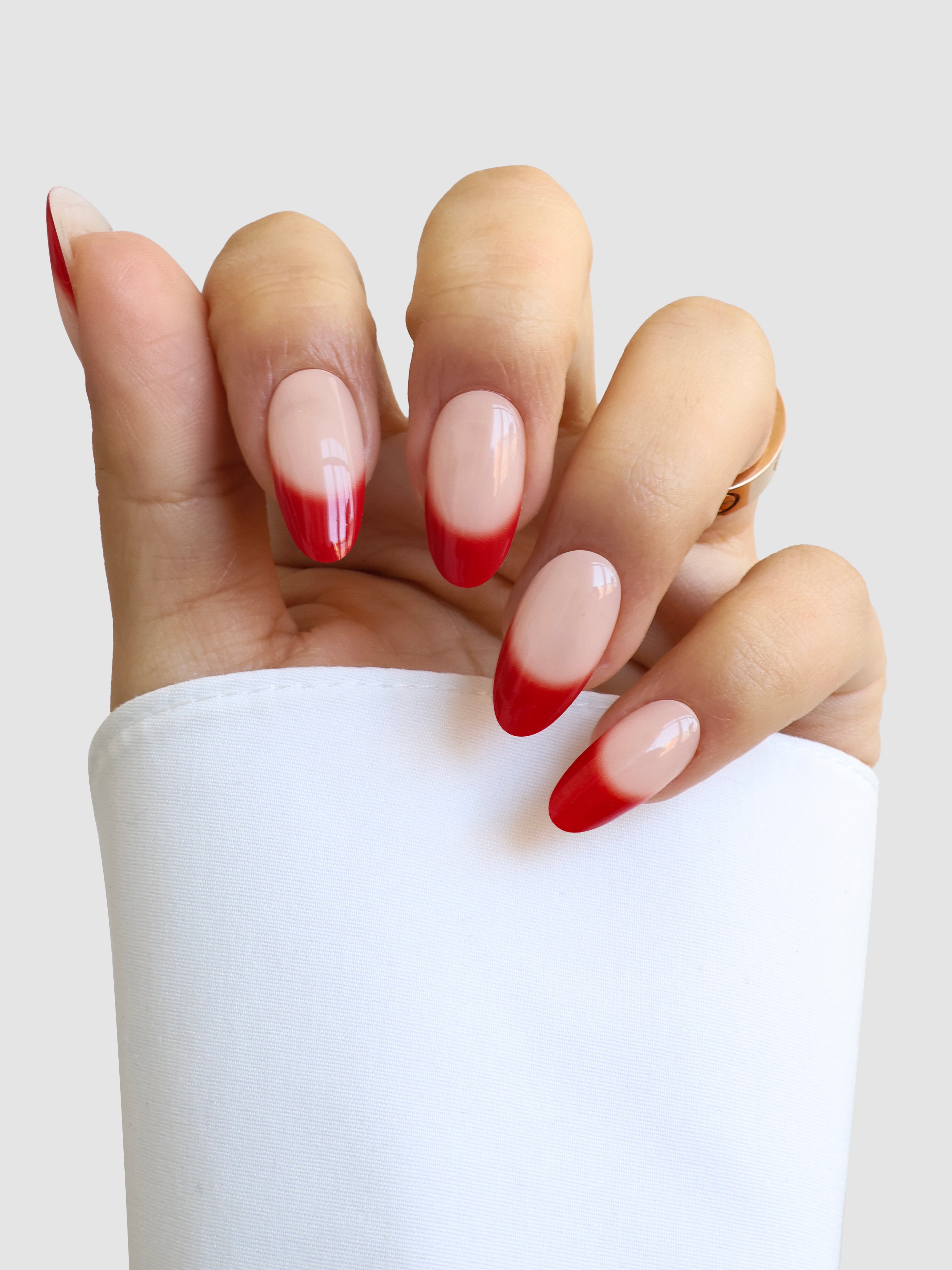 A hand with almond-shaped nails sporting Sassy Saints' Madame Rouge Gel Wrap—nude nails with bold red tips—is held upright against a white background, and the person is dressed in a white long-sleeved top.