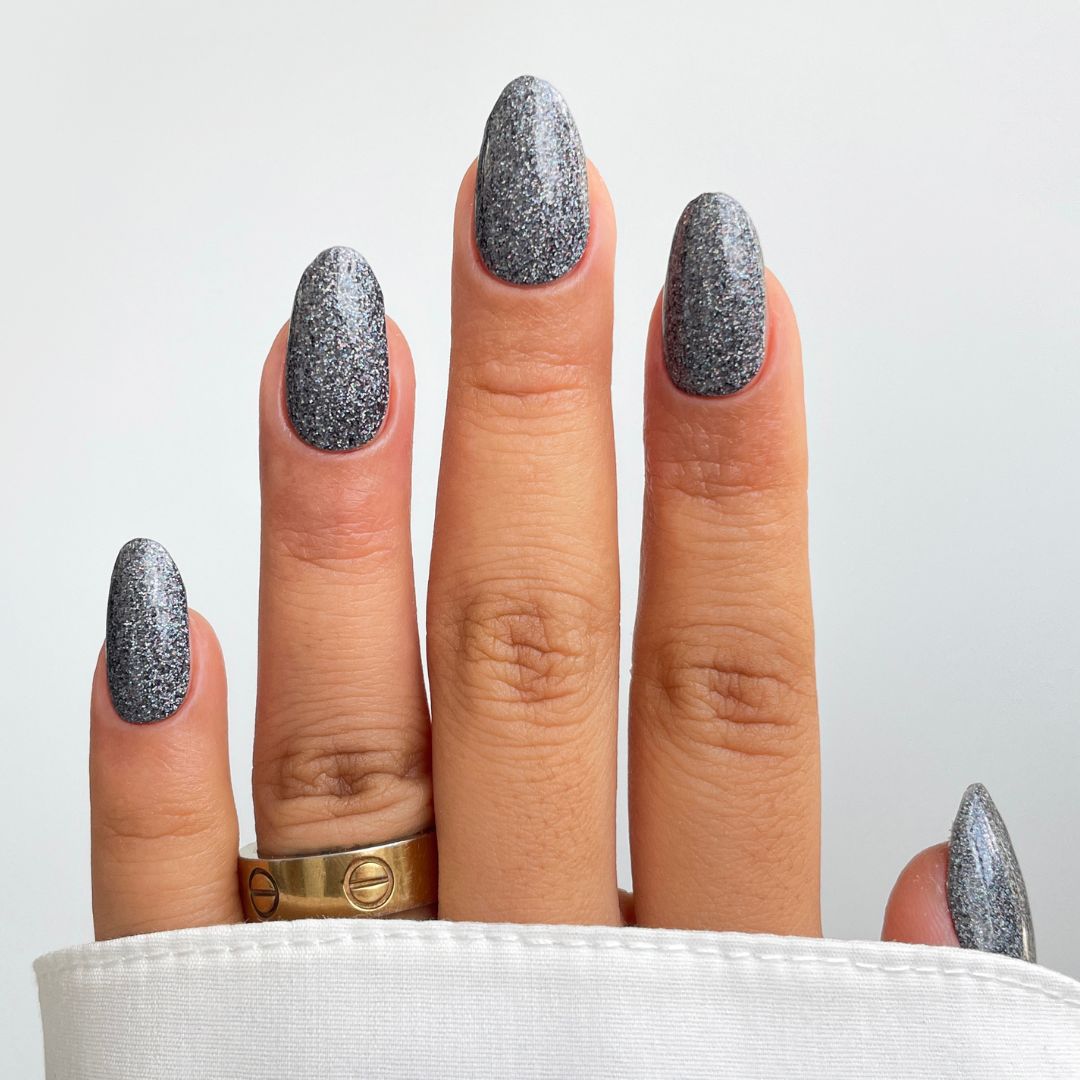 A hand showcases Sassy Saints - Brings The Salon Home’s Midnight Glimmer Gel Wrap on oval nails, paired with two gold rings on the middle finger and a white cuffed shirt, set against a plain white background.