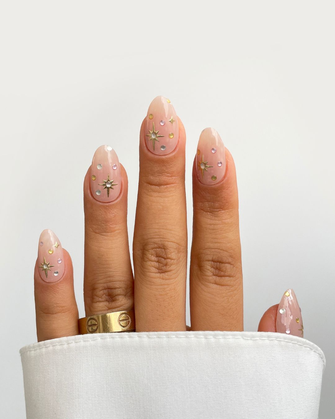 A hand features Milkyway Gel Wrap by Sassy Saints - Brings The Salon Home: almond-shaped, nude nails with pastel gems and metallic stars. A gold ring and a white sleeve partially cover the hand.