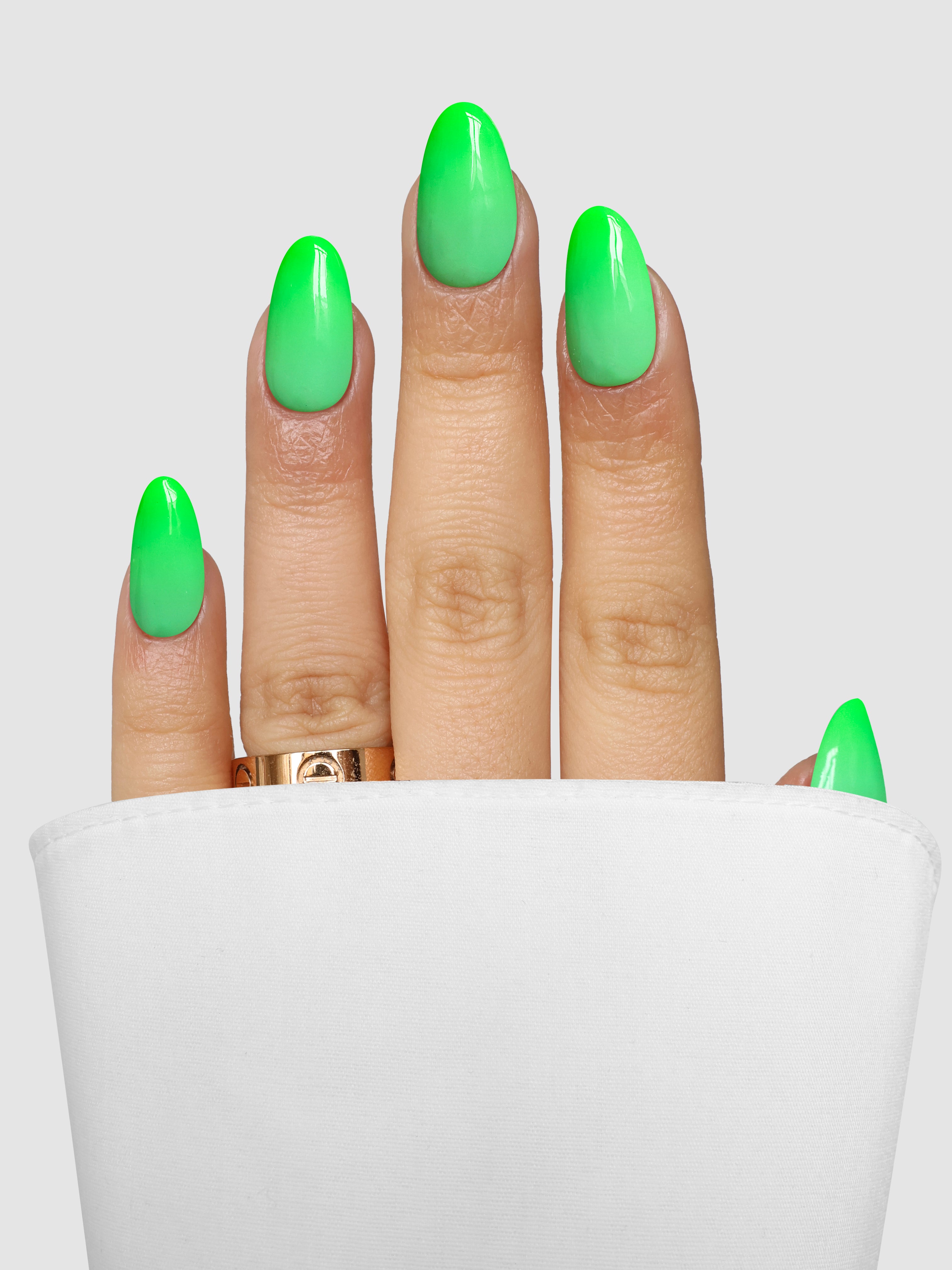 A hand with nails in a neon green ombre—created using Sassy Saints Mint Magic Dipping Powder—peeks from a white sleeve. The person wears a gold ring on their middle finger against a plain gray background.