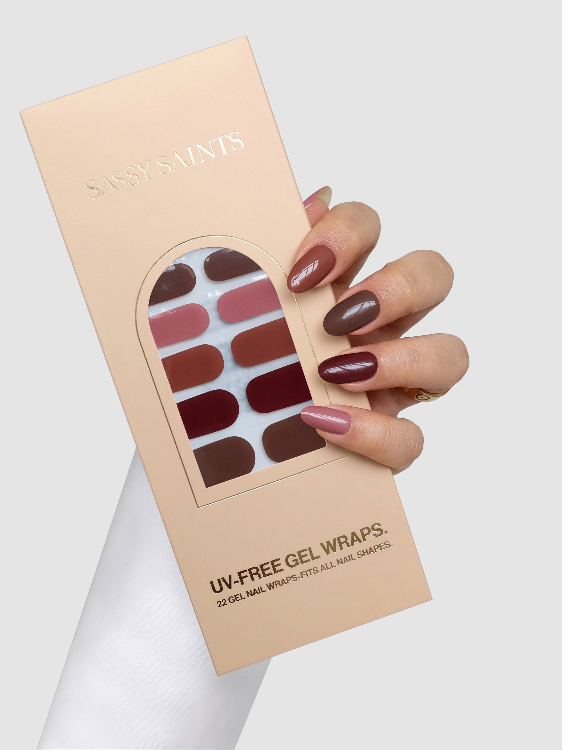 A hand with painted nails holds a box of Sassy Saints – Brings The Salon Home Mocha Melt Gel Wraps. The gel wraps display elegant shades of red and pink. The person wears a white sleeve and a gold ring.