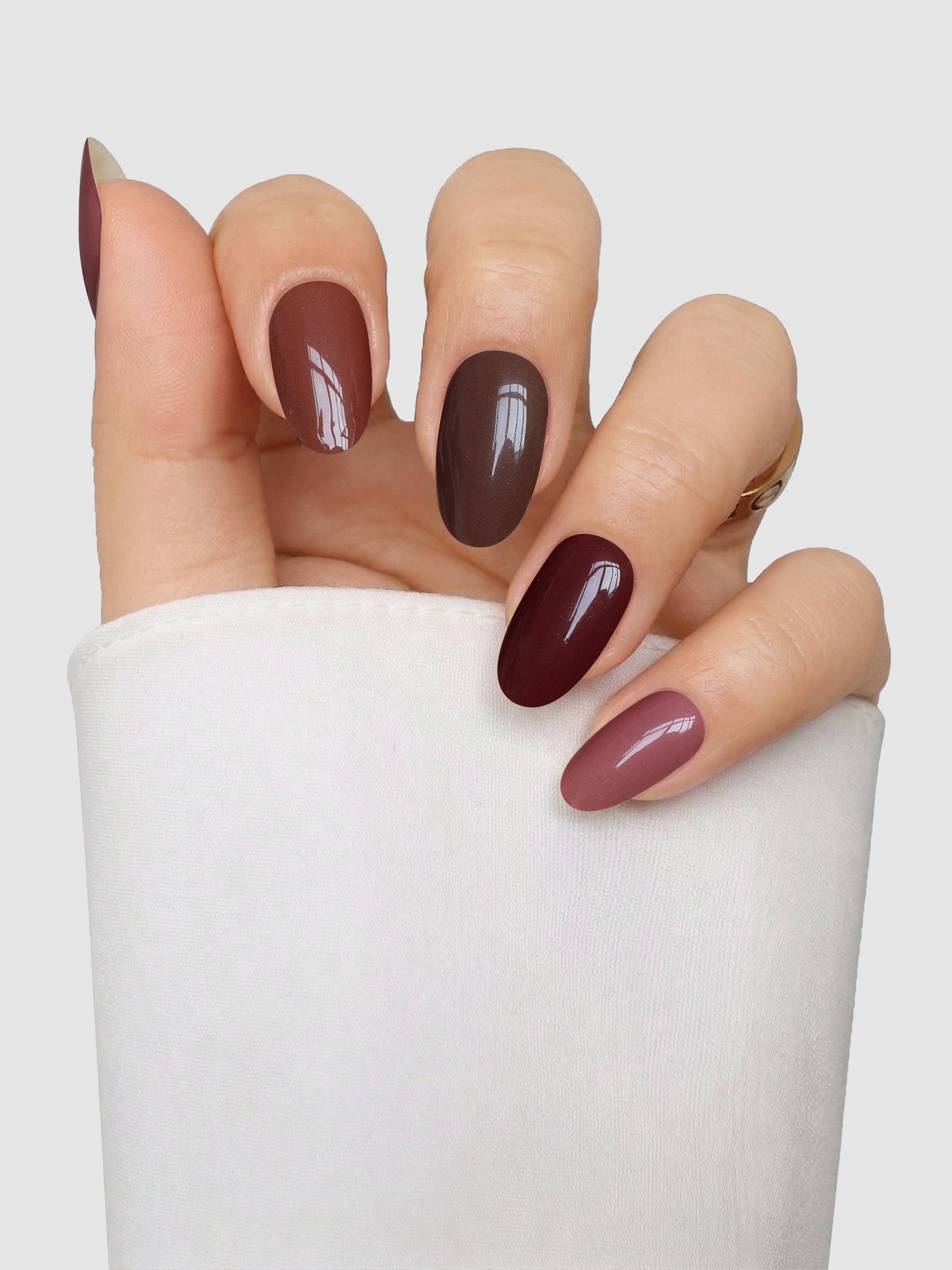 A hand with almond-shaped nails featuring Sassy Saints - Brings The Salon Home’s Mocha Melt Gel Wrap in various brown and mauve tones, held against a light gray background. The person is dressed in a long-sleeve white top.