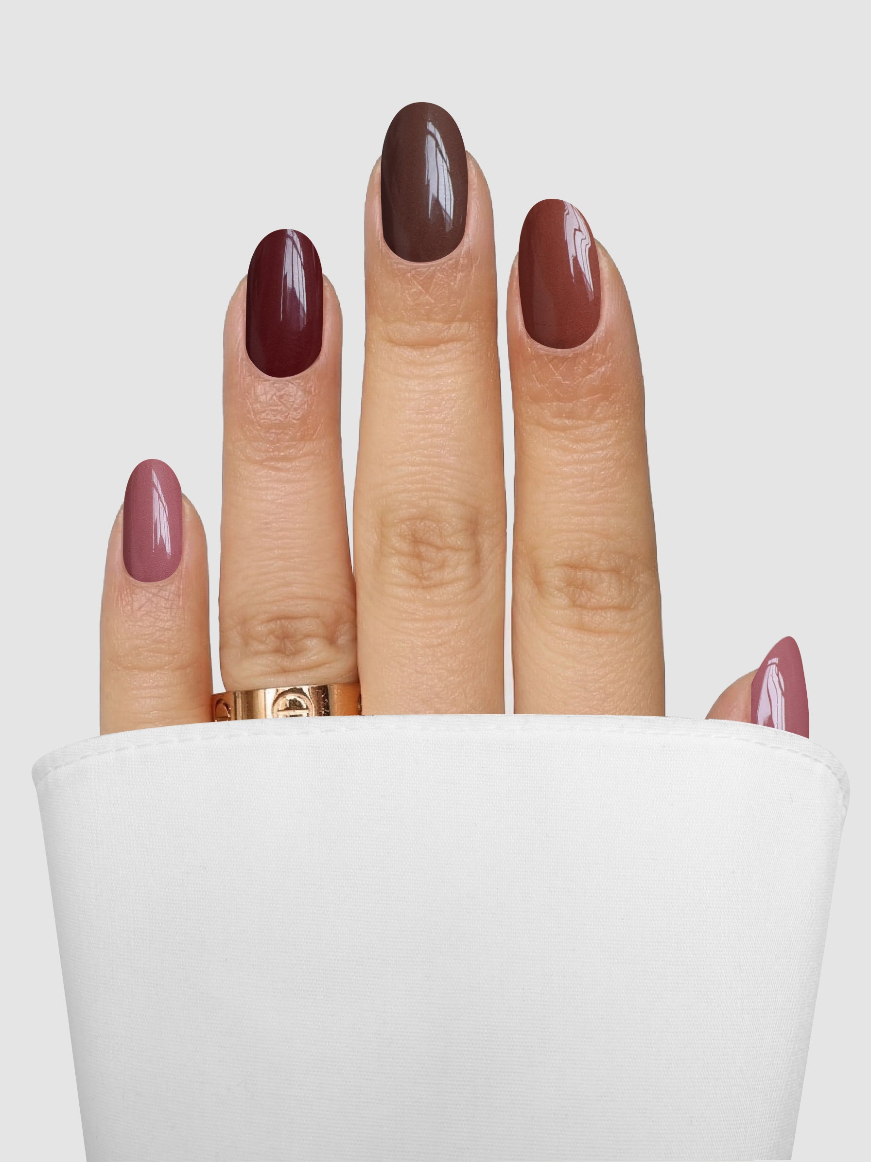 A hand adorned with Sassy Saints - Brings The Salon Home’s Mocha Melt Gel Wrap nail polish, featuring a gradient from pink to brown, rests against a white sleeve, highlighted by a gold ring on the middle finger.