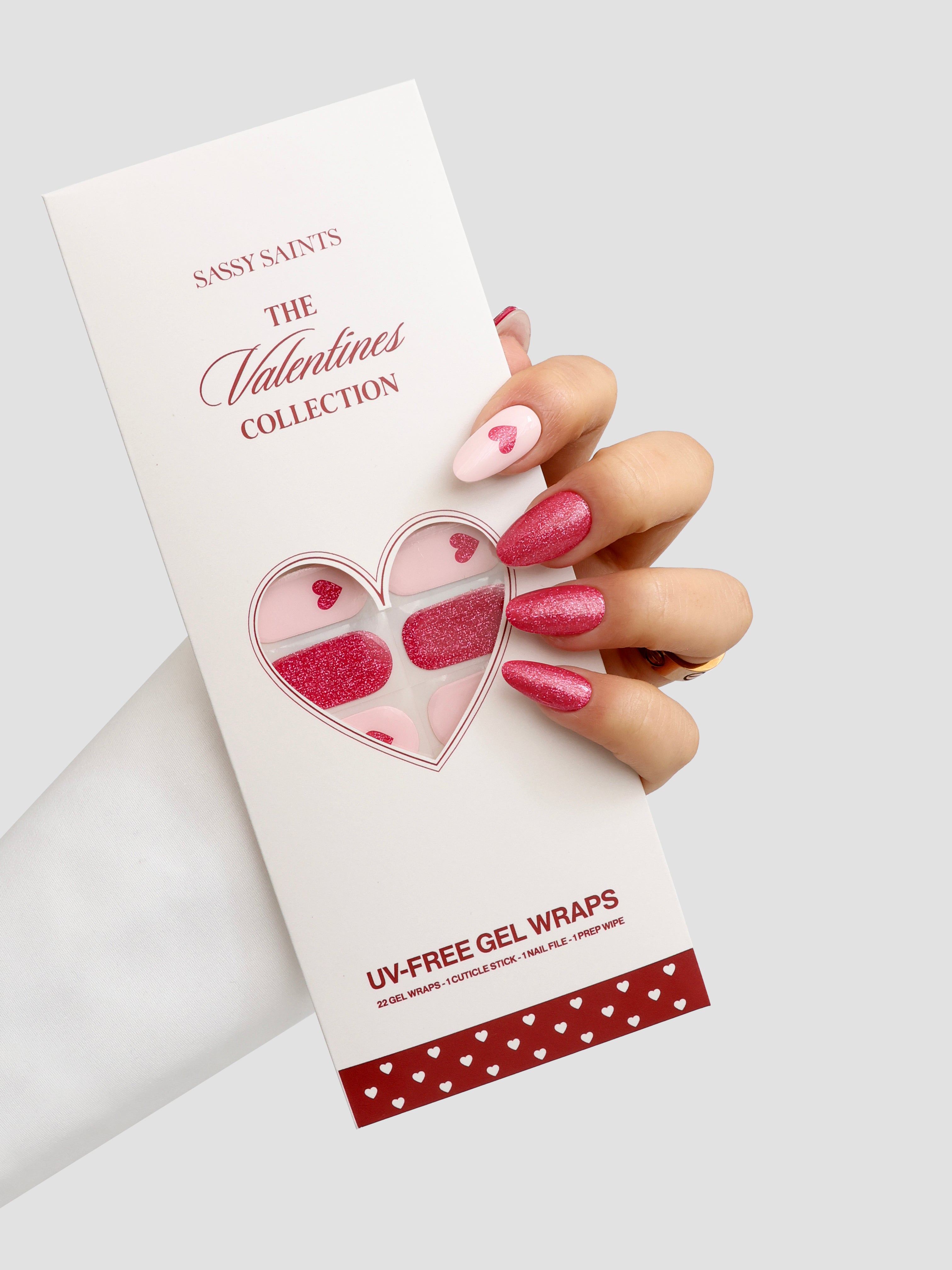 A hand with pink glitter and white heart-themed nail wraps holds a Sassy Saints - Brings The Salon Home Modern Romance Gel Wrap package, featuring a heart-shaped window that displays the nail wraps inside.