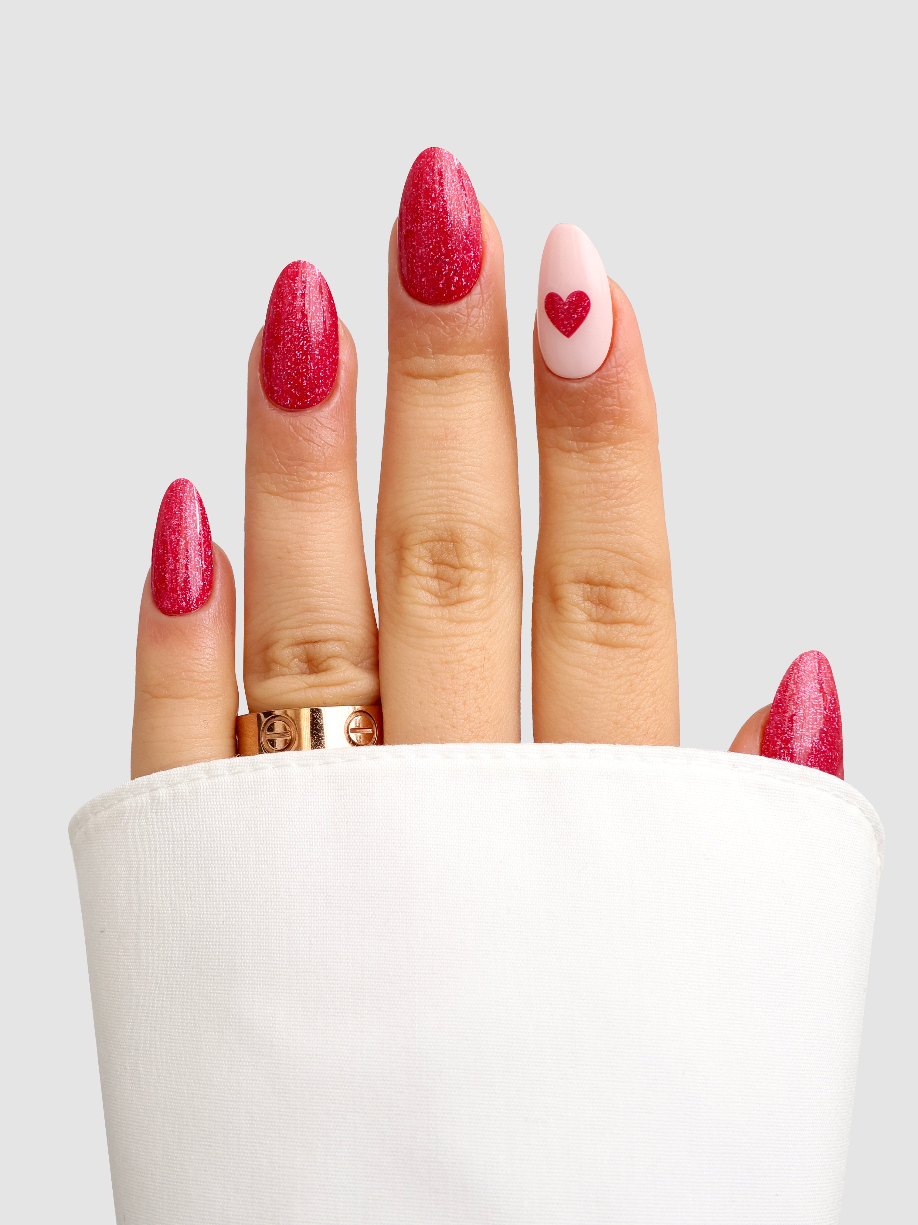 A hand models Sassy Saints' Modern Romance Gel Wrap: pink glittery nails with a red heart accent, paired with a gold ring and partly covered by a white sleeve, shown against a gray background.