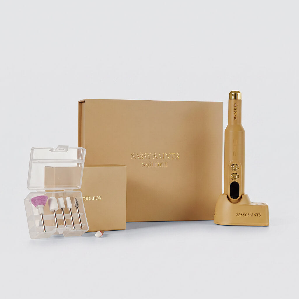 A gold Sassy Saints At-Home E-File with attachments, a gold box labeled “Sassy Saints - Brings The Salon Home,” a smaller accessory box, and a clear case filled with assorted nail drill bits—all crafted for professional-quality nail care—are displayed on a white background. This Sassy Saints At-Home E-File set makes it easy to achieve salon-quality results from the comfort of home.