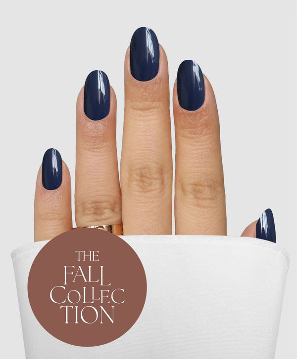 A hand with nails polished in Sassy Saints - Brings The Salon Home’s Slate State Gel Wrap is shown against a light background. The fingers are partly covered by a white cloth, and a brown circle features “THE FALL COLLECTION” in white text.