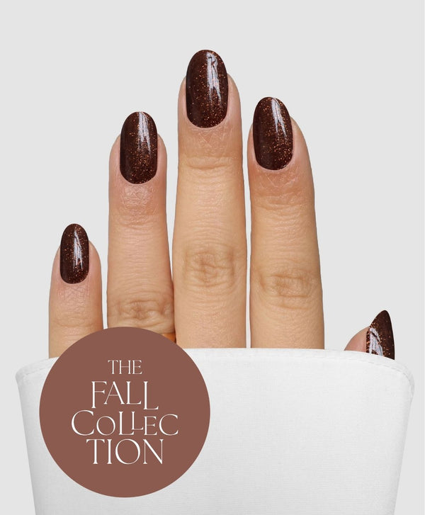 A hand with short, almond-shaped nails is adorned with Sassy Saints - Brings The Salon Home’s Bronze Behaviour Gel Wrap, featuring a glittery dark brown finish, and rests against a white background. A brown circle displays “THE FALL COLLECTION” in white text.