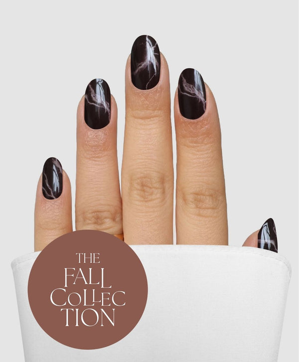 A hand showcasing dark, glossy, marble-patterned nails—created with the Truffle Swirl Gel Wrap by Sassy Saints - Brings The Salon Home—peeks out from a white sleeve. In the bottom left corner, a brown circle displays “THE FALL COLLECTION” in white text.
